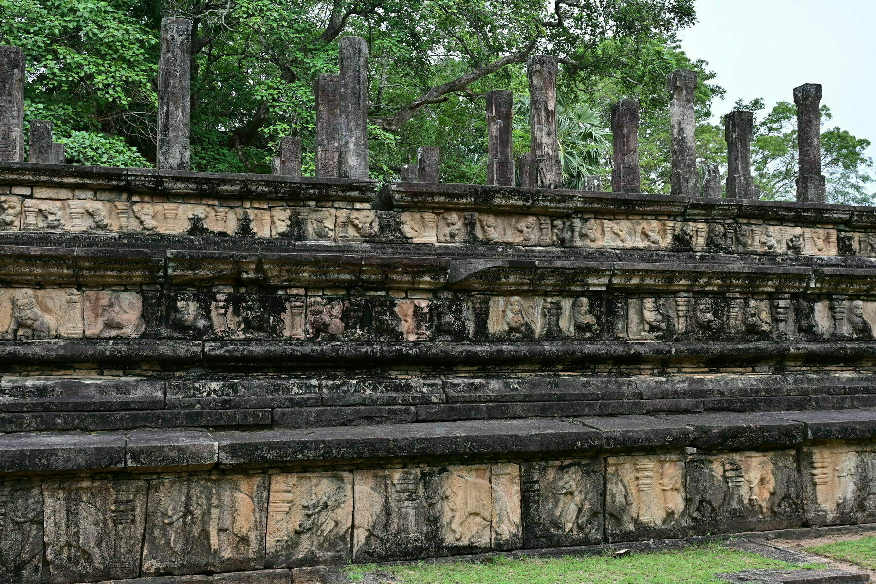 Auto-generated description: Ancient stone carvings adorn a tiered, weathered wall surrounded by lush greenery.