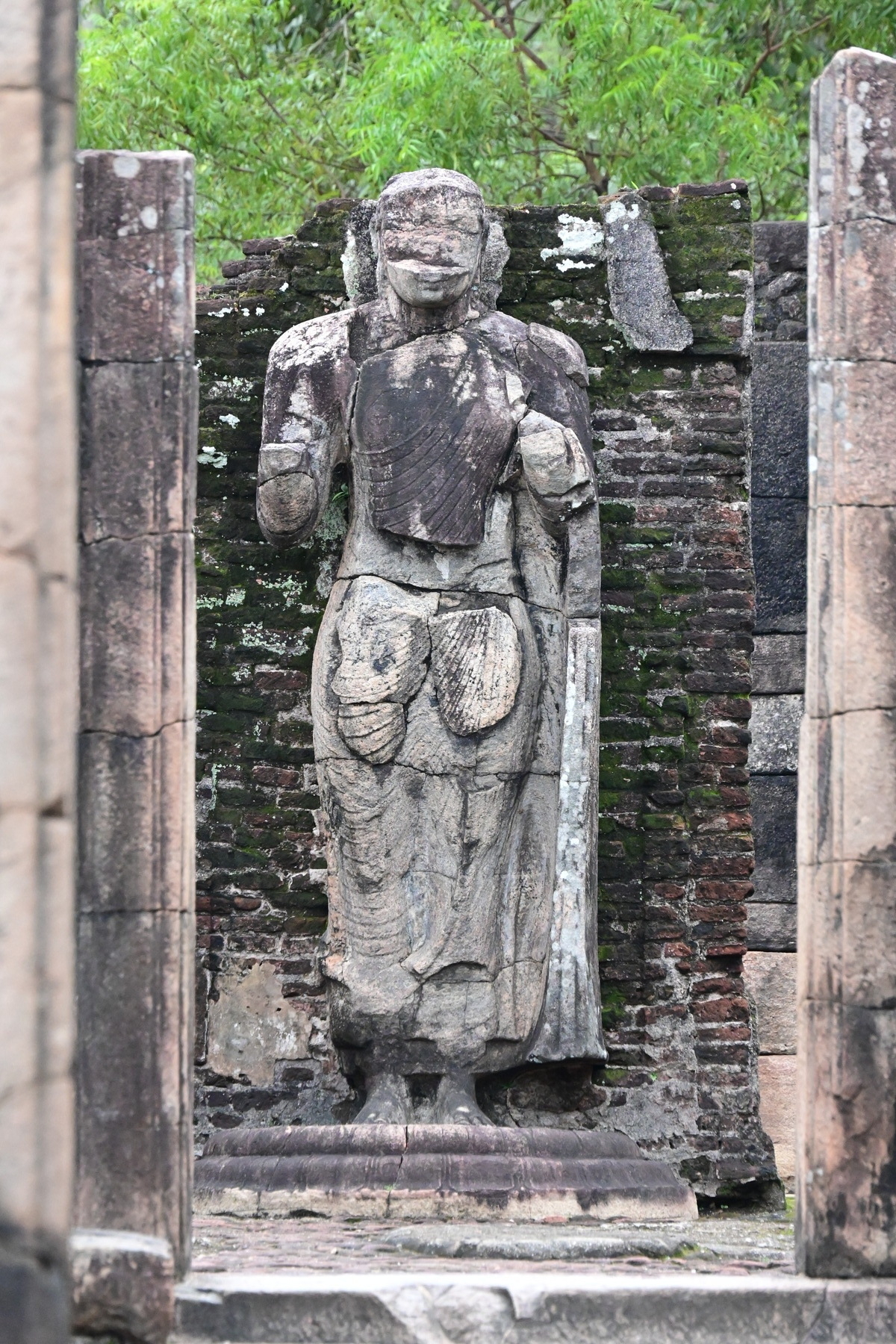 Auto-generated description: An ancient stone statue stands amidst historical ruins and greenery.