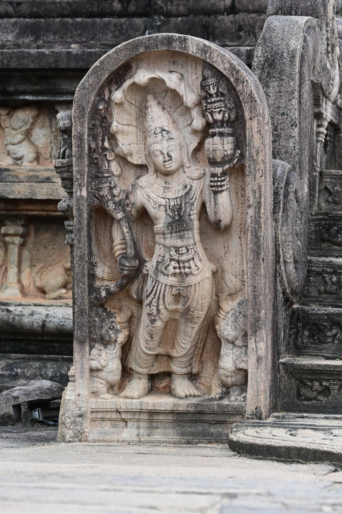 Auto-generated description: An intricately carved ancient stone statue depicts a figure holding a staff or club and a conch shell, standing in an ornate archway.