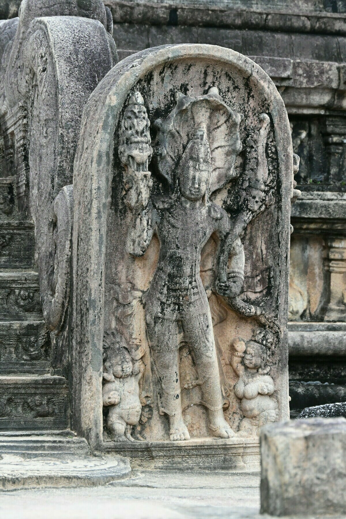 Auto-generated description: A stone carving depicts a standing figure in an elaborate, traditional attire with intricate details, flanked by smaller figures.