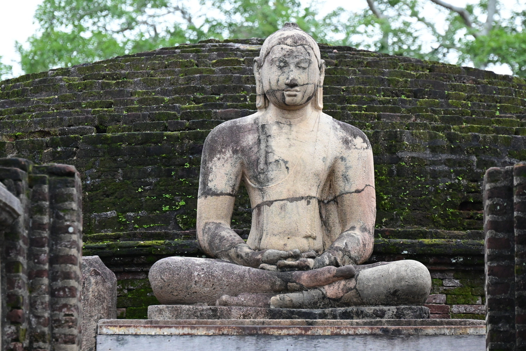 Auto-generated description: A seated stone statue of a serene figure is set against a backdrop of ancient, moss-covered brickwork and greenery.