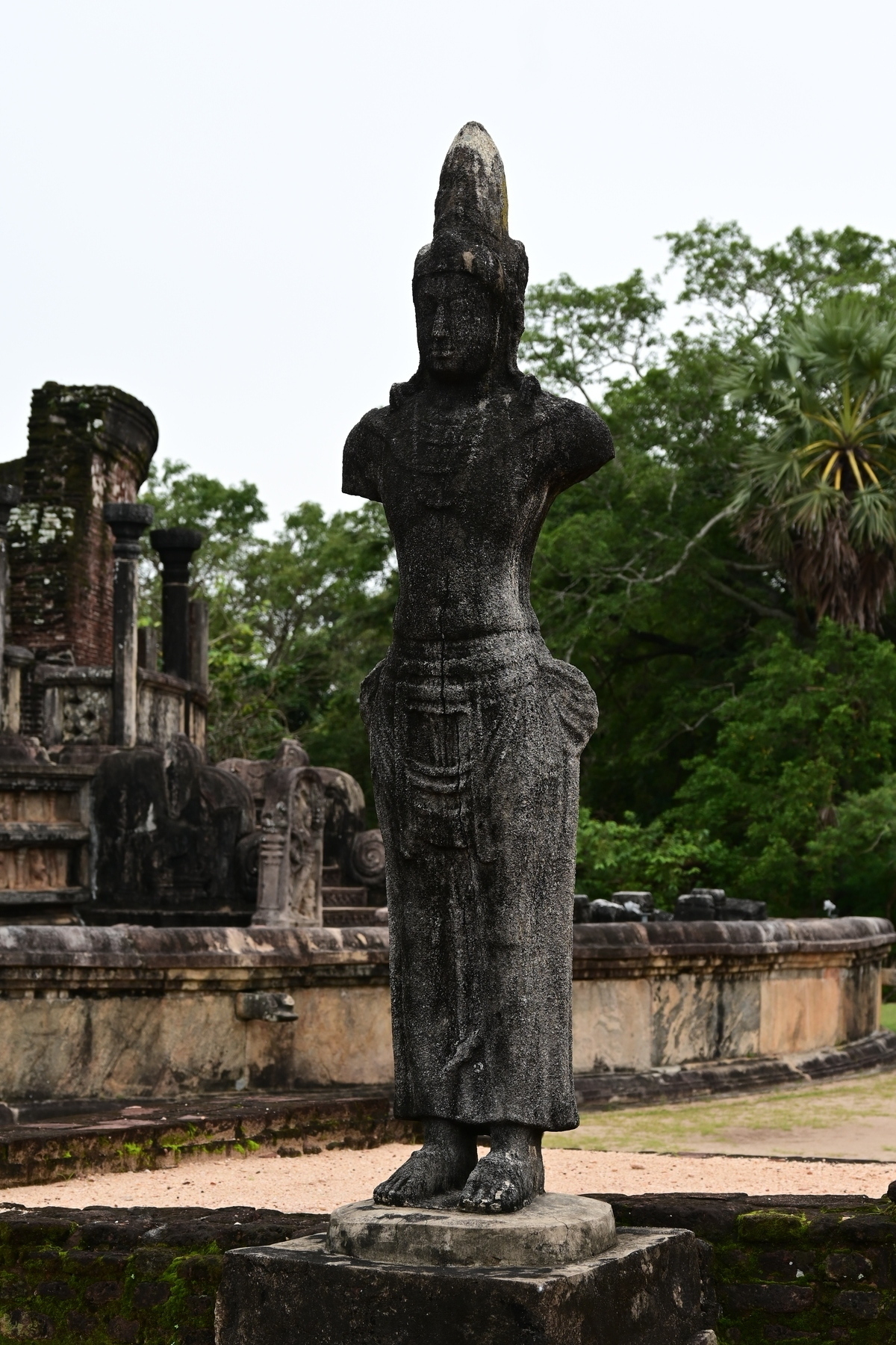 Auto-generated description: A stone statue stands in front of ancient ruins surrounded by greenery.