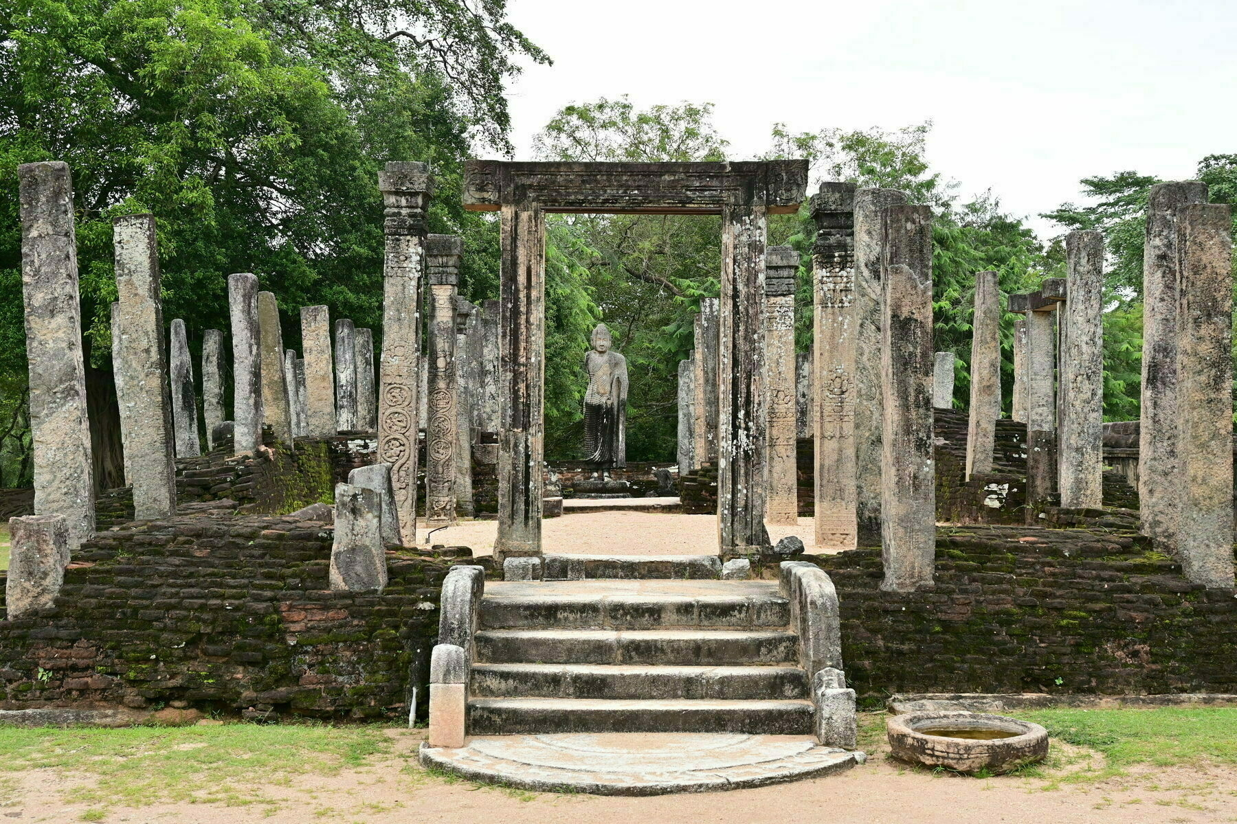 Auto-generated description: Ancient stone pillars and steps lead to a statue standing within a historic ruin surrounded by lush greenery.