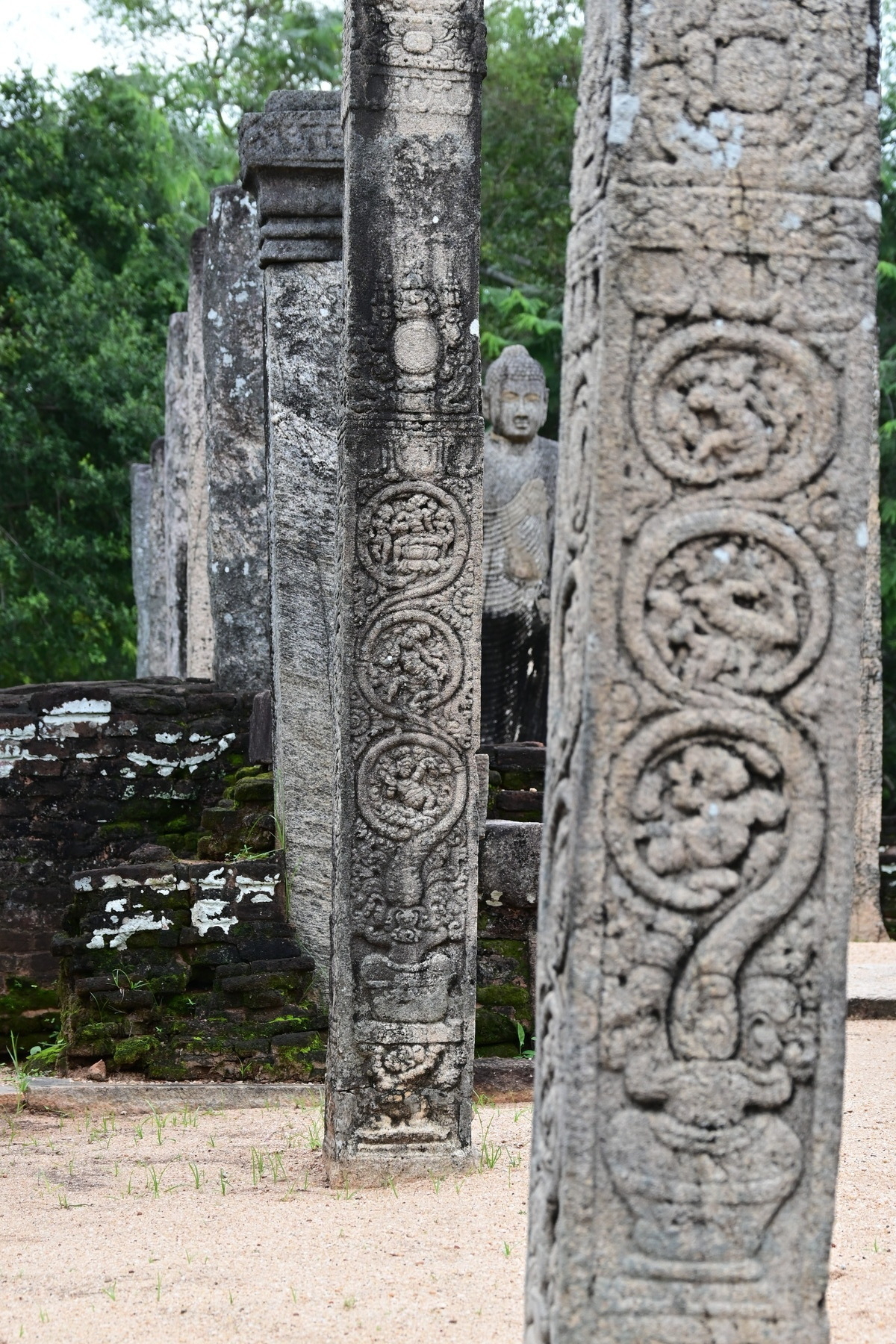 Auto-generated description: Ancient stone pillars with intricate carvings stand in front of a seated statue amid a lush green backdrop.