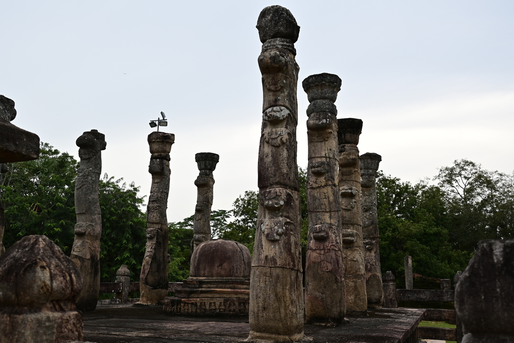 Auto-generated description: Ancient stone pillars with intricate carvings stand amidst lush greenery under a cloudy sky.