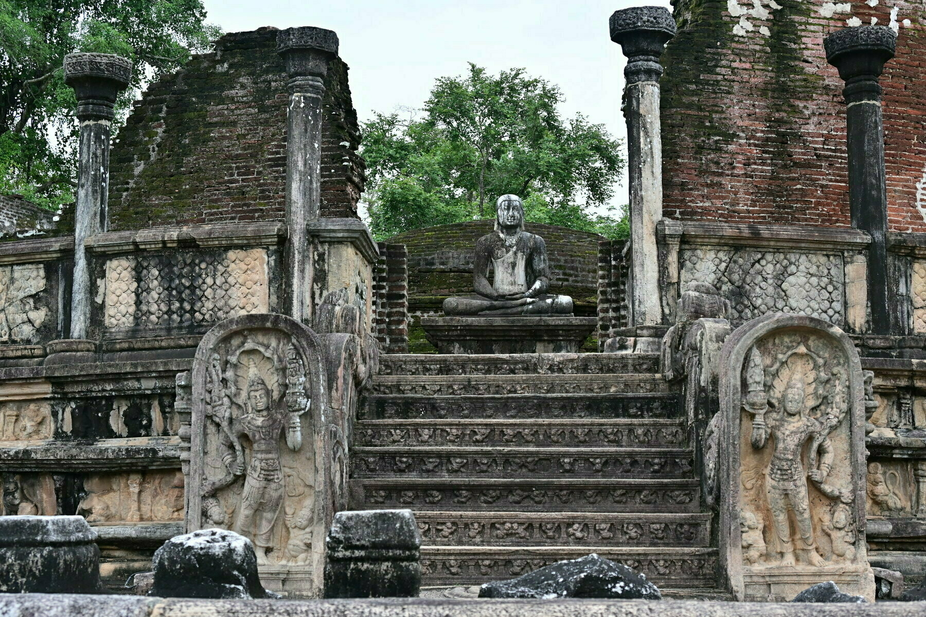 Auto-generated description: A seated Buddha statue is surrounded by intricate stone carvings and flanked by ancient, weathered pillars on a raised platform.