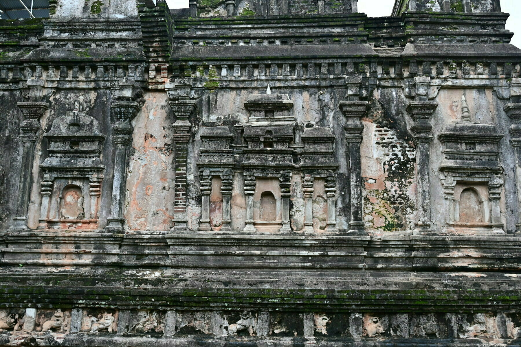 Auto-generated description: An ancient, weathered stone wall features intricate carvings and layered architectural details, partially covered in moss.