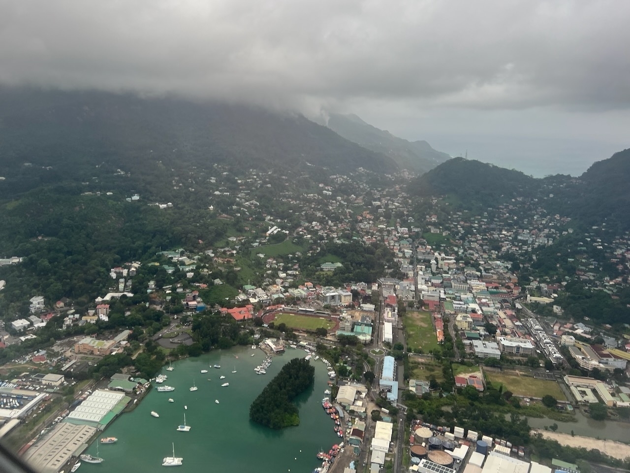 Auto-generated description: Aerial view of a cityscape surrounded by lush green hills and cloudy skies, with a marina filled with boats.