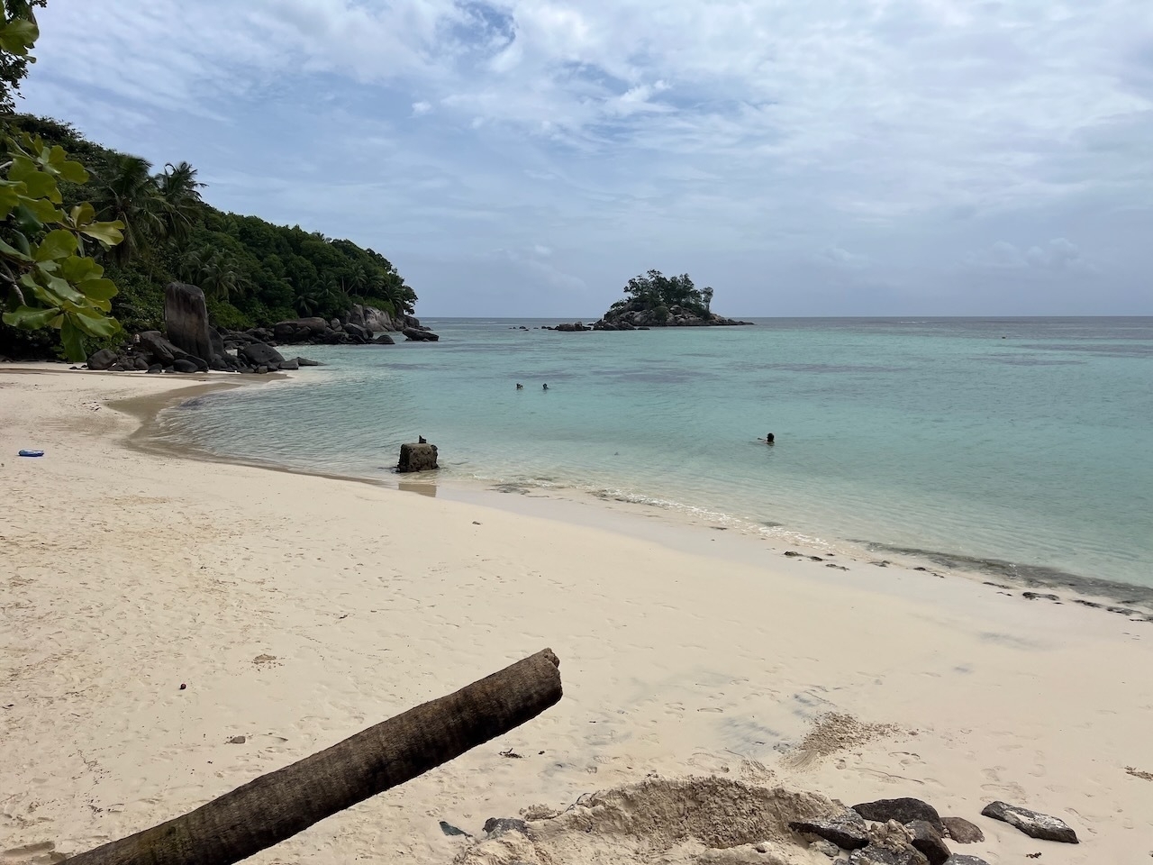 Auto-generated description: A serene beach scene features white sand, calm turquoise water, a few swimmers, and a small island in the distance.