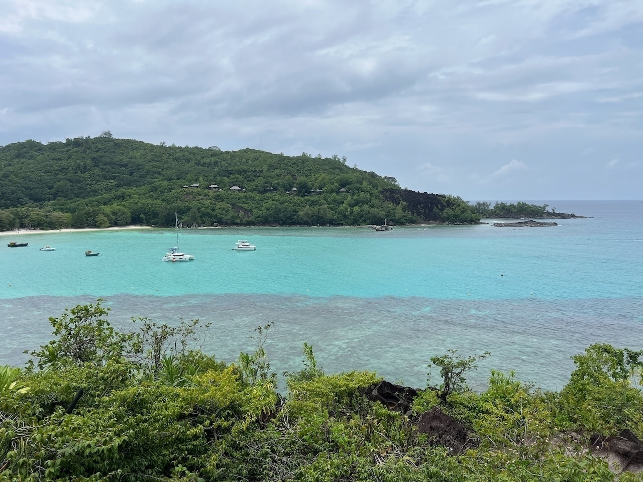 Auto-generated description: A scenic view showcases a tranquil turquoise bay with several boats, surrounded by lush, green hills under a partly cloudy sky.