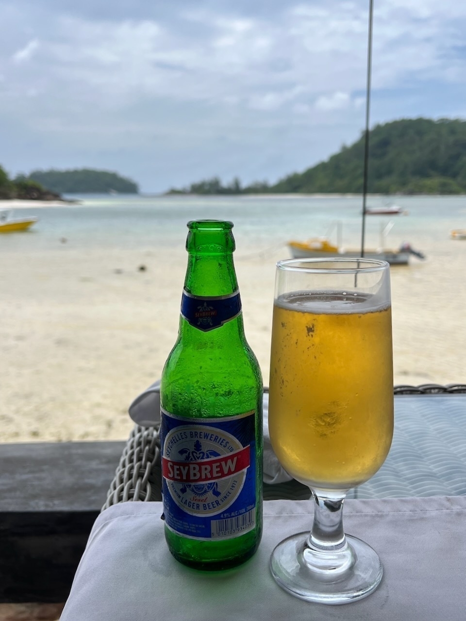 Auto-generated description: A bottle and glass of Seybrew beer are on a table overlooking a tranquil beach with a view of boats and green hills in the background.