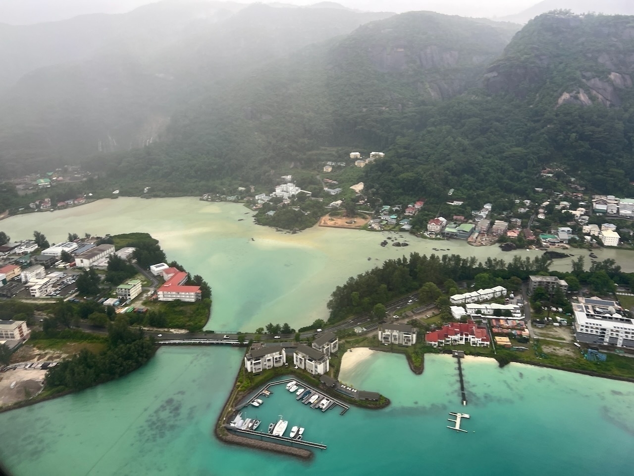 Auto-generated description: An aerial view of a coastal town features lush greenery, houses, and a marina with turquoise waters.