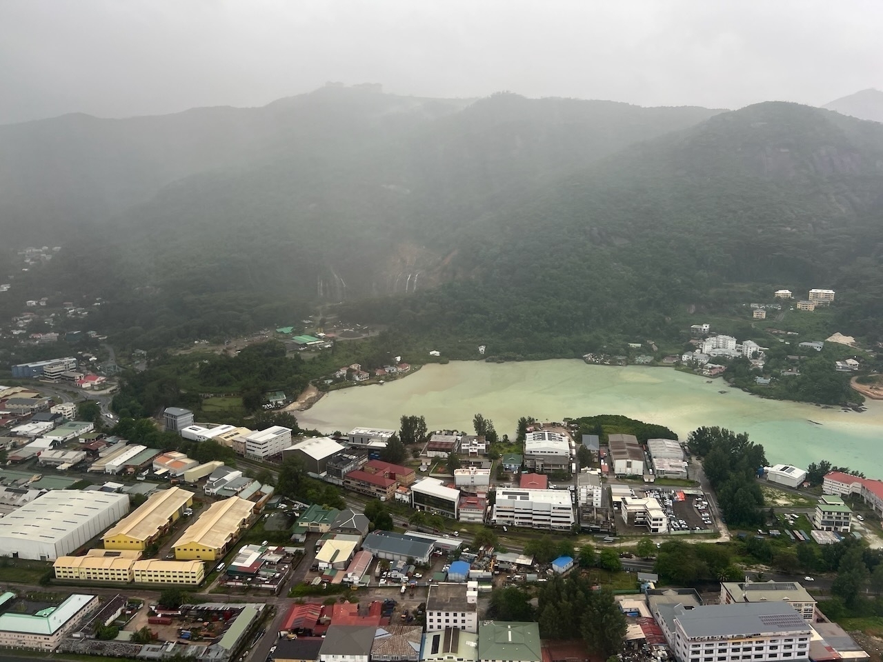 Auto-generated description: Aerial view of a coastal town with buildings, lush green hills, and a cloudy sky.