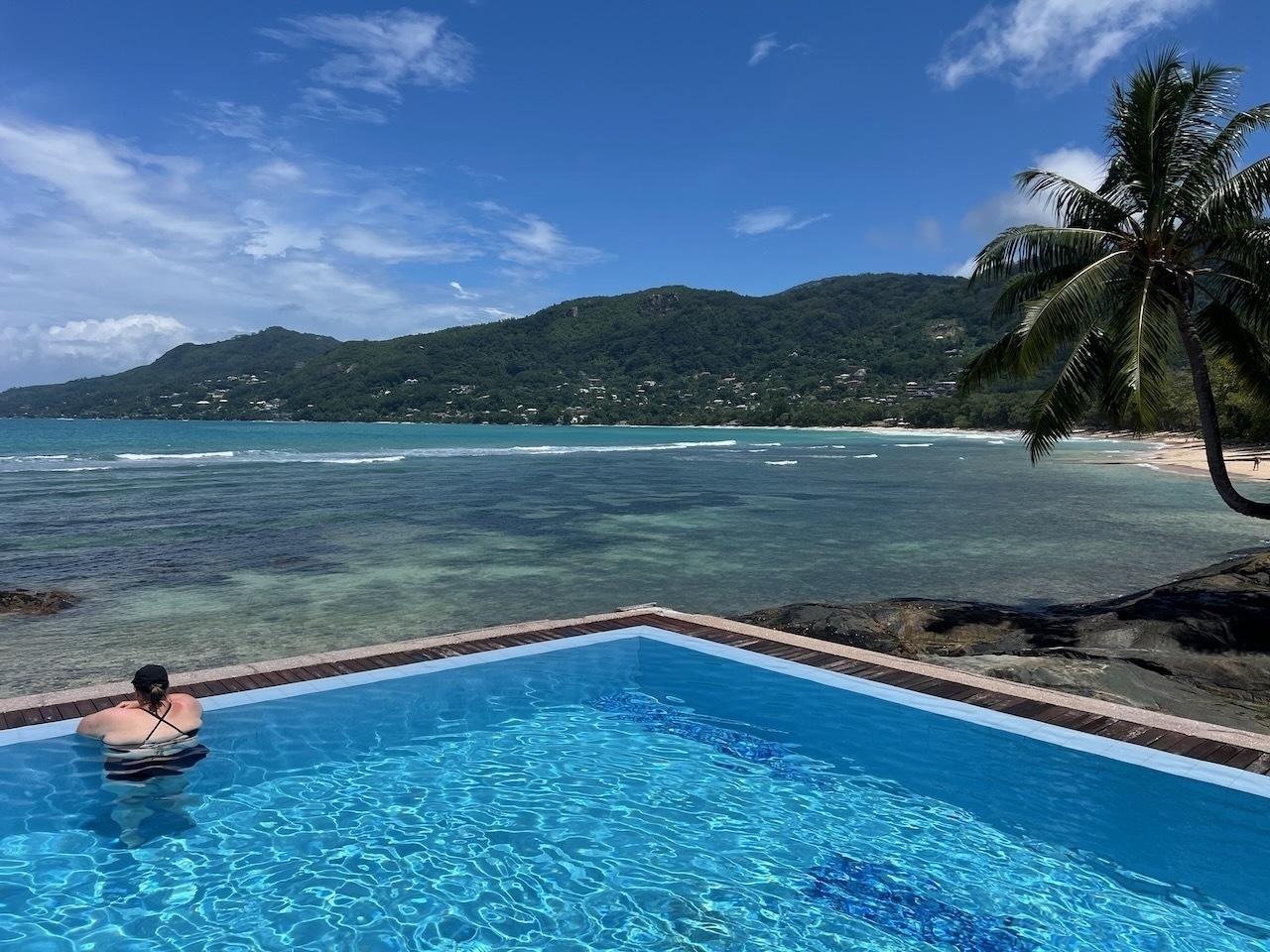 Auto-generated description: A person is relaxing at the edge of a pool, overlooking a scenic ocean view with mountains and palm trees in the background.