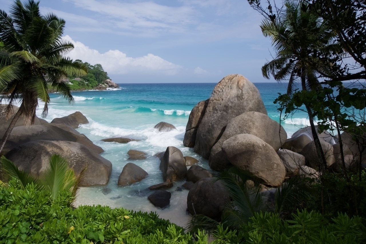 Auto-generated description: A serene tropical beach scene features large boulders, turquoise waves, and lush green foliage under a partly cloudy sky.