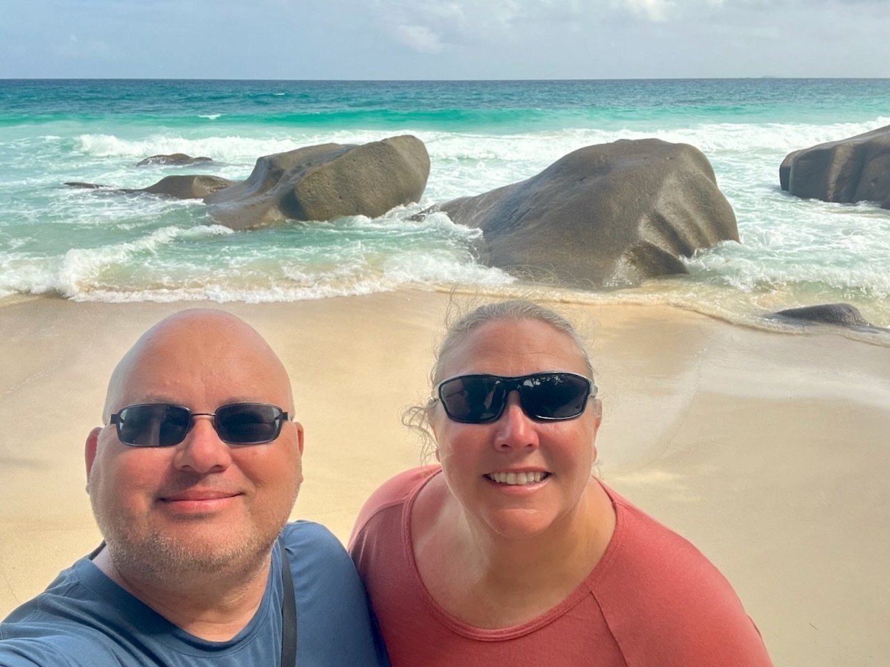 Auto-generated description: Two people wearing sunglasses are smiling on a beach with waves and large rocks in the background.