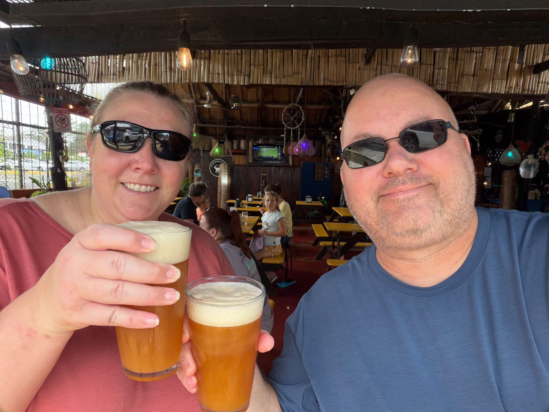Two people wearing sunglasses are smiling and holding pint glasses of beer in an outdoor setting with wooden decor.
