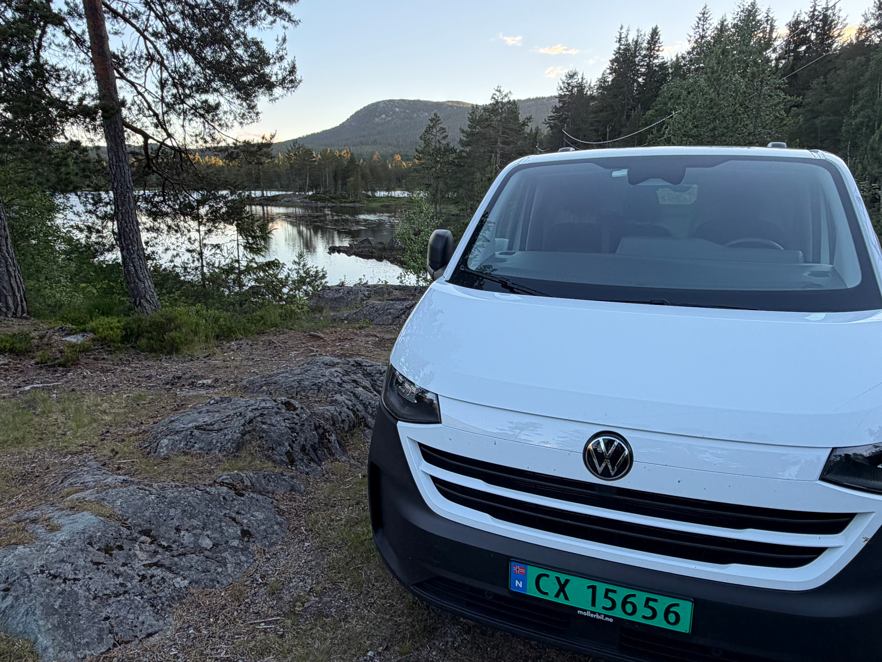 A white Volkswagen van is parked on rocky terrain near a calm lake surrounded by trees and hills.