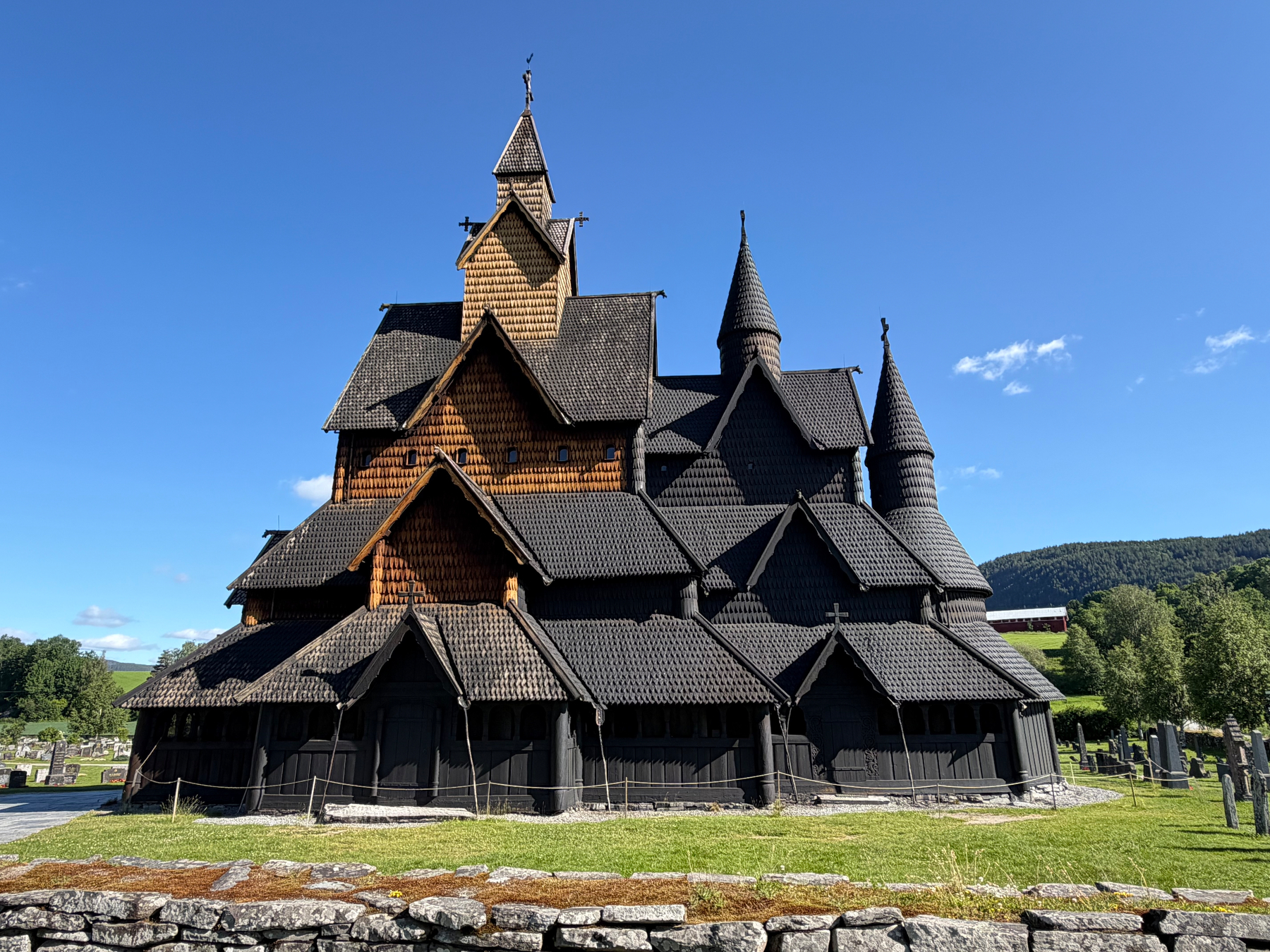 A large, intricately designed stave church with multiple steep, pointed roofs stands under a clear blue sky surrounded by a grassy area and a cemetery.
