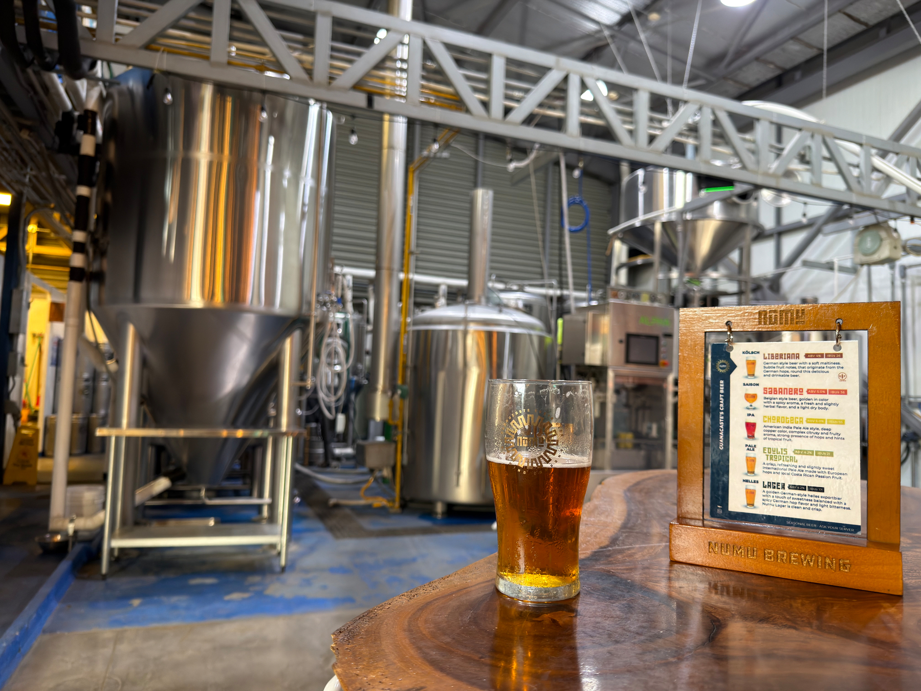 A pint of beer sits on a wooden table inside a brewery, with brewing equipment and a menu on display in the background.