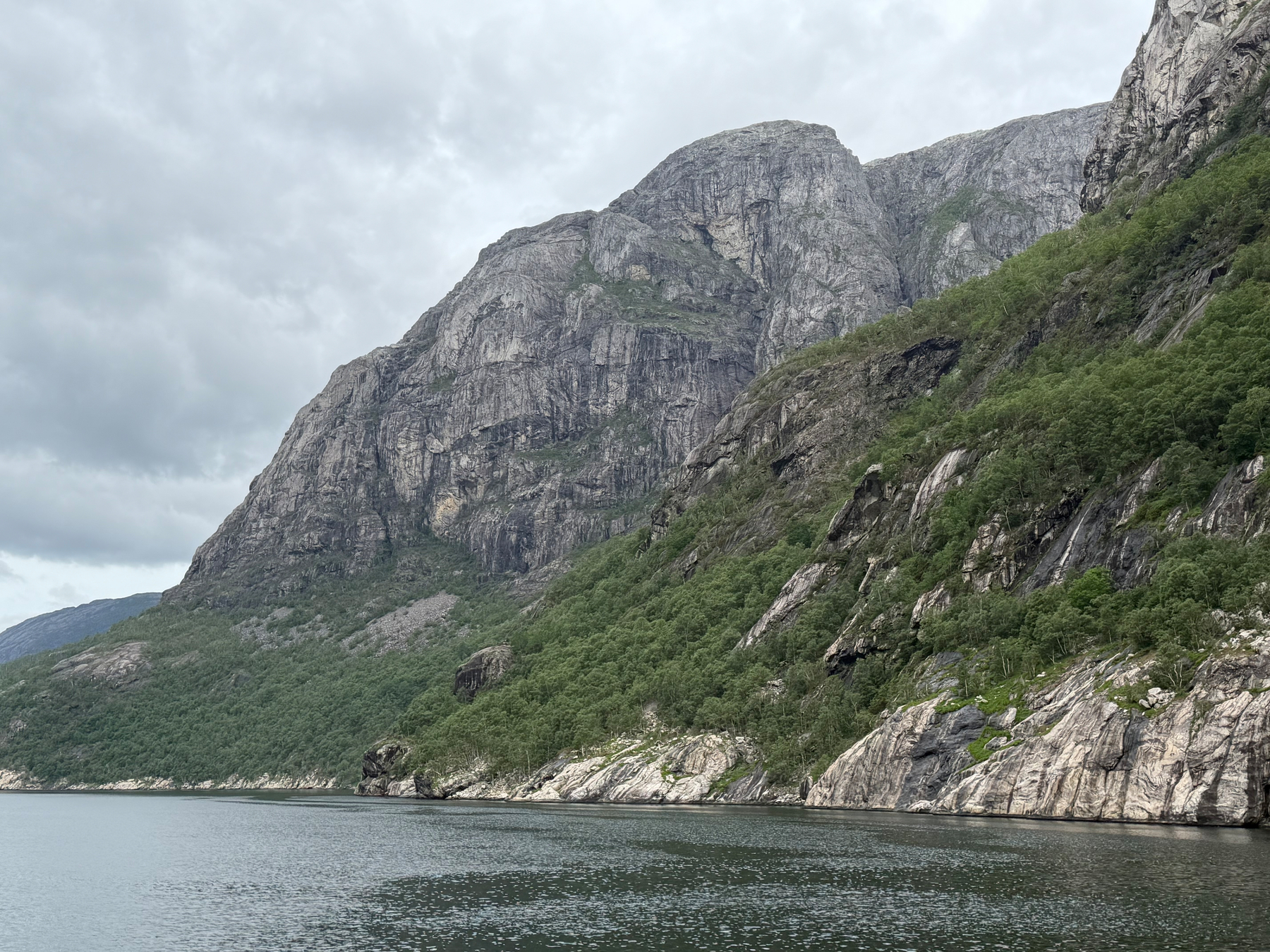 A serene body of water lies at the base of towering rocky cliffs covered with patches of greenery under a cloudy sky.