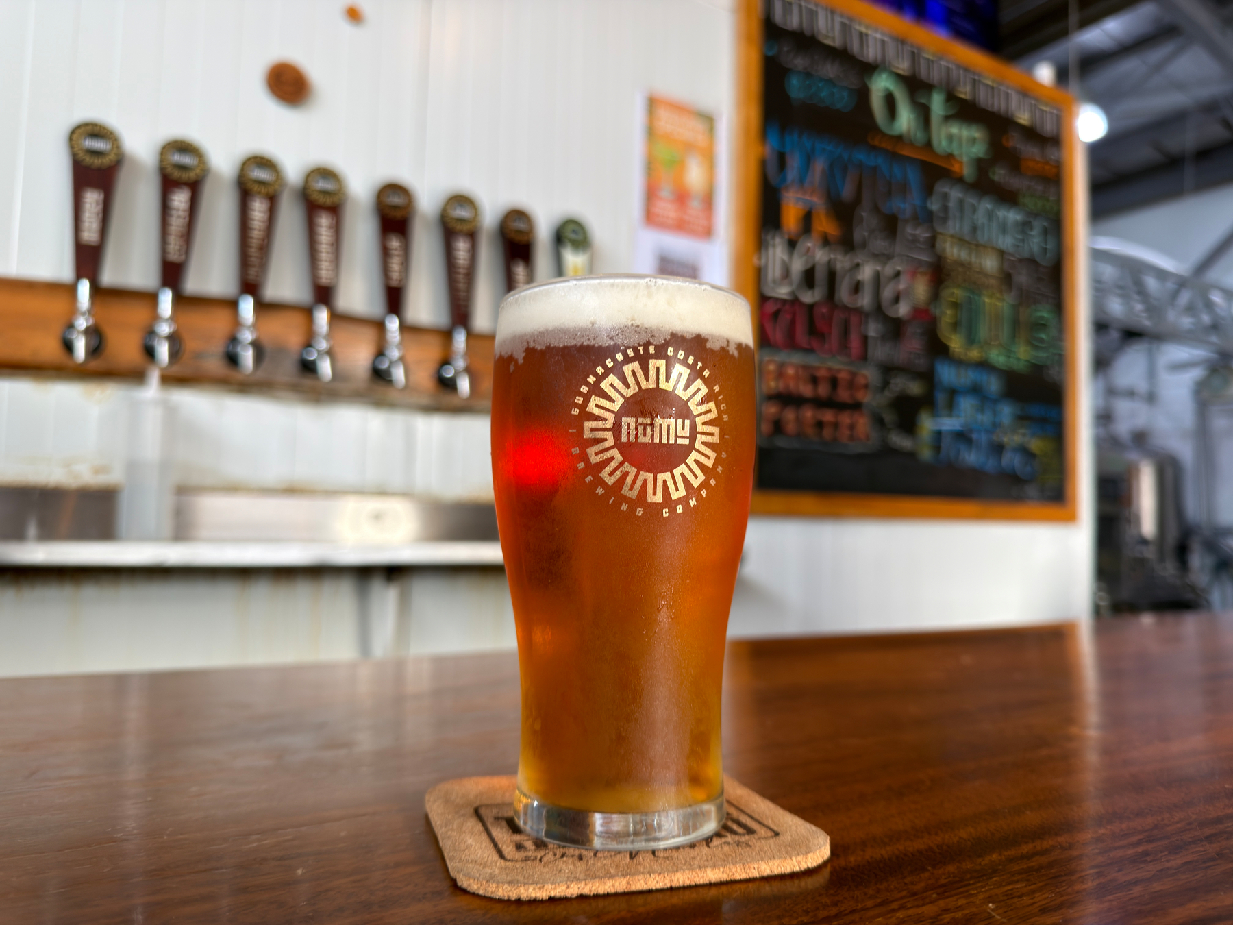 A glass of beer with a foamy head is placed on a wooden bar top in front of a row of taps and a colorful chalkboard menu.
