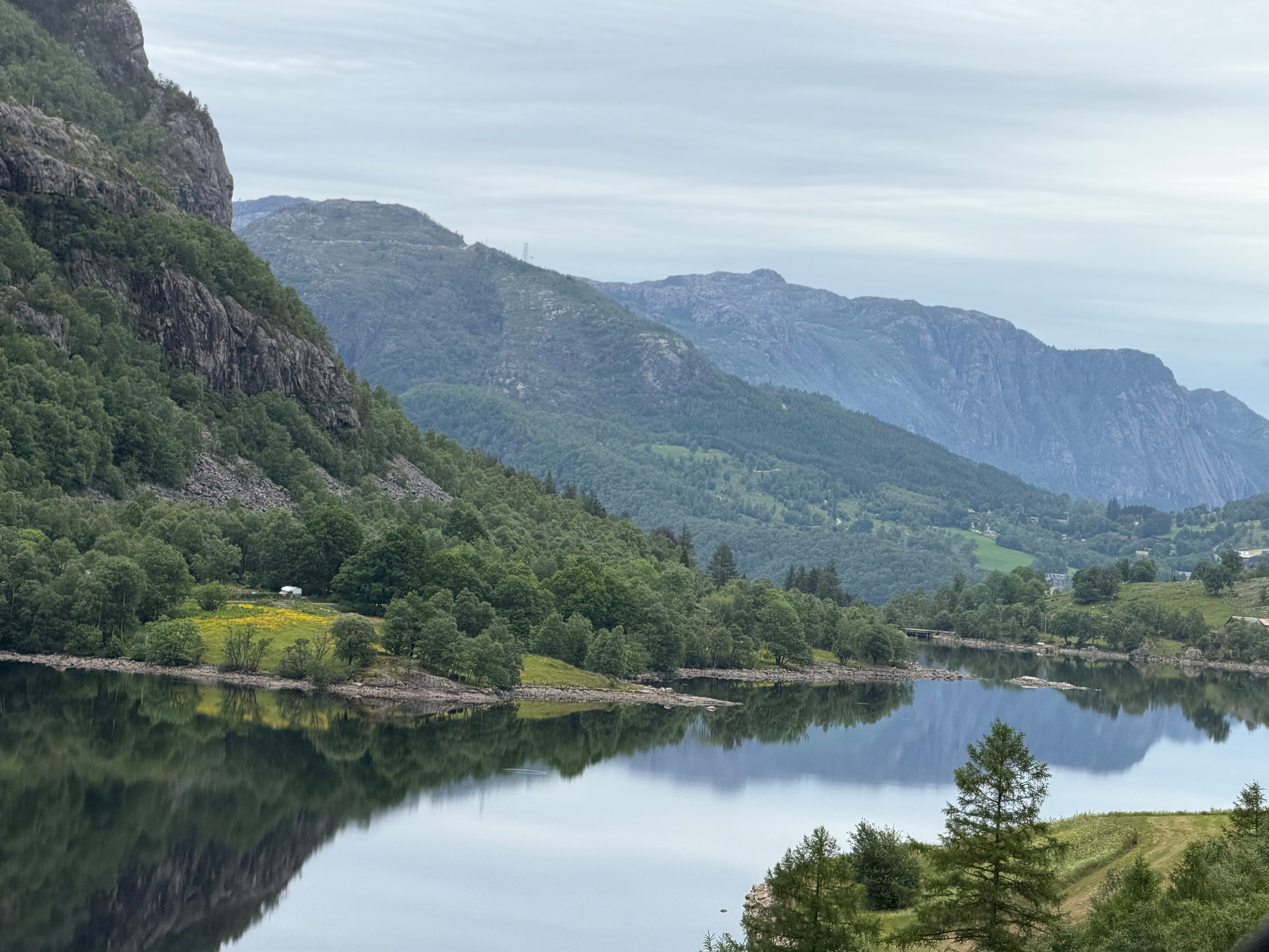 A serene lake reflects the surrounding lush green hills and distant mountains under a cloudy sky.