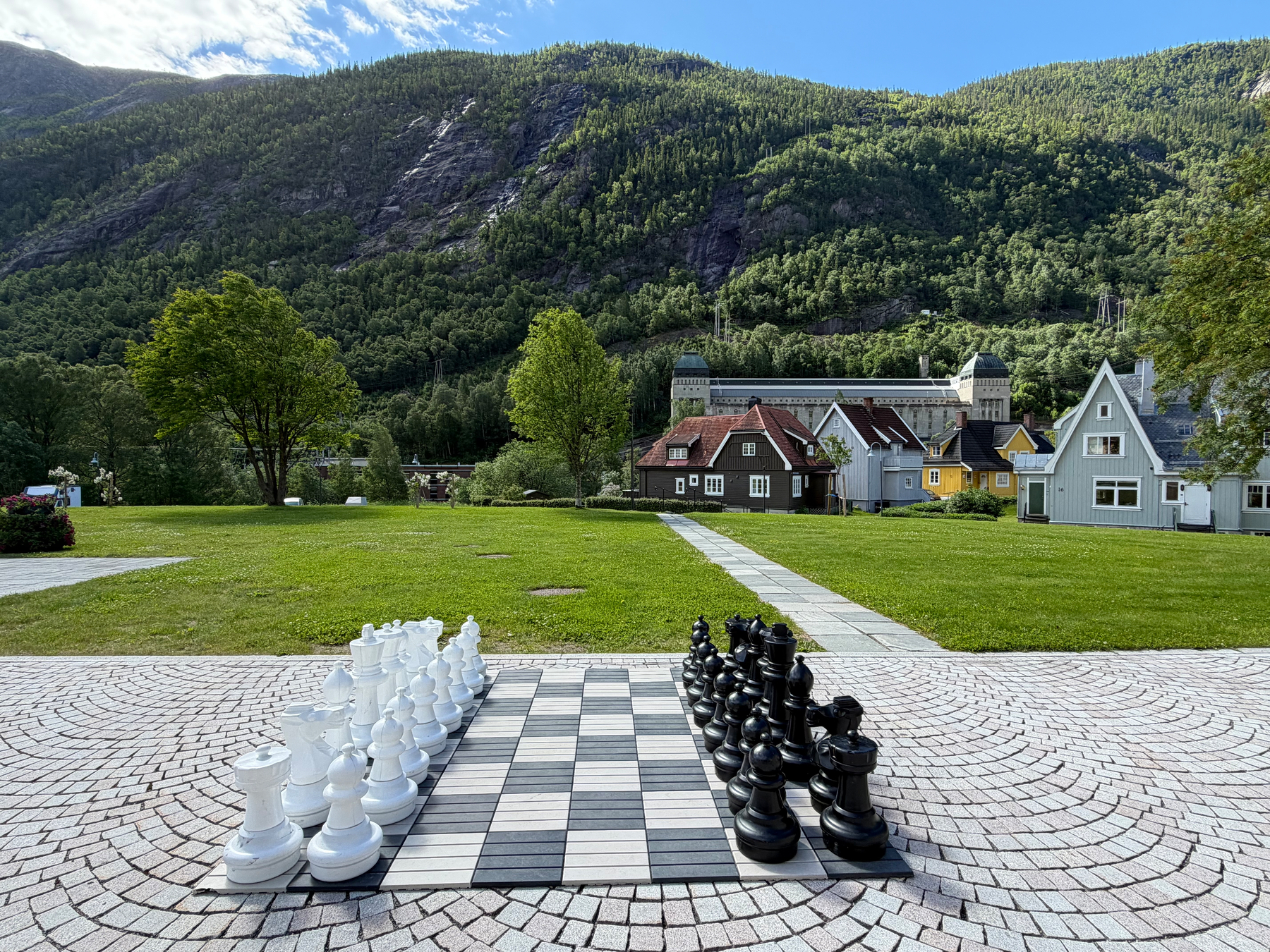 A large outdoor chessboard with oversized pieces is set up on a tiled patio overlooking a grassy area, quaint houses, and forested mountains under a partly cloudy sky.