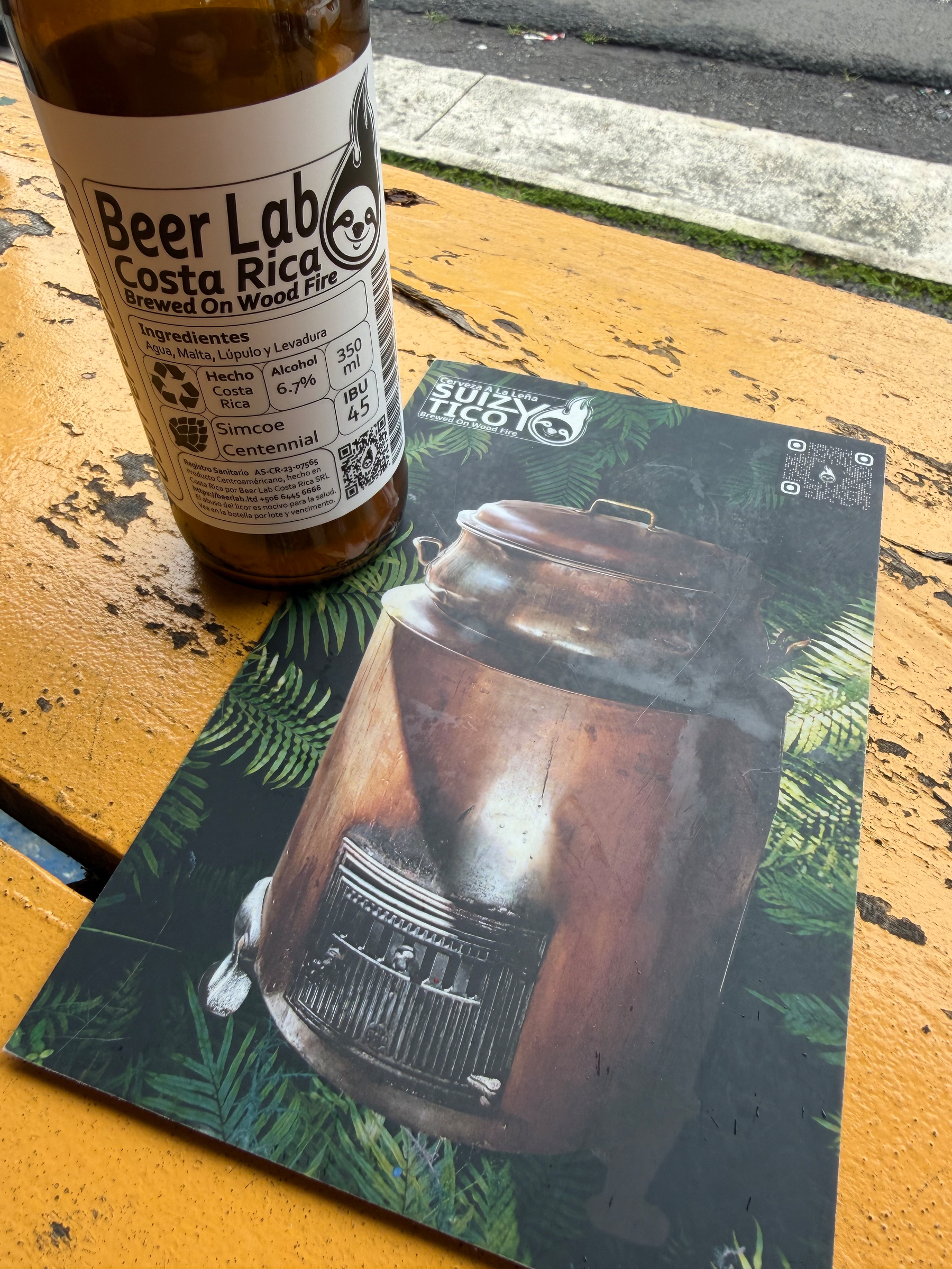 A bottle of Beer Lab Costa Rica stands on a yellow wooden surface next to an artistic beer menu featuring an image of a small wood-burning stove.