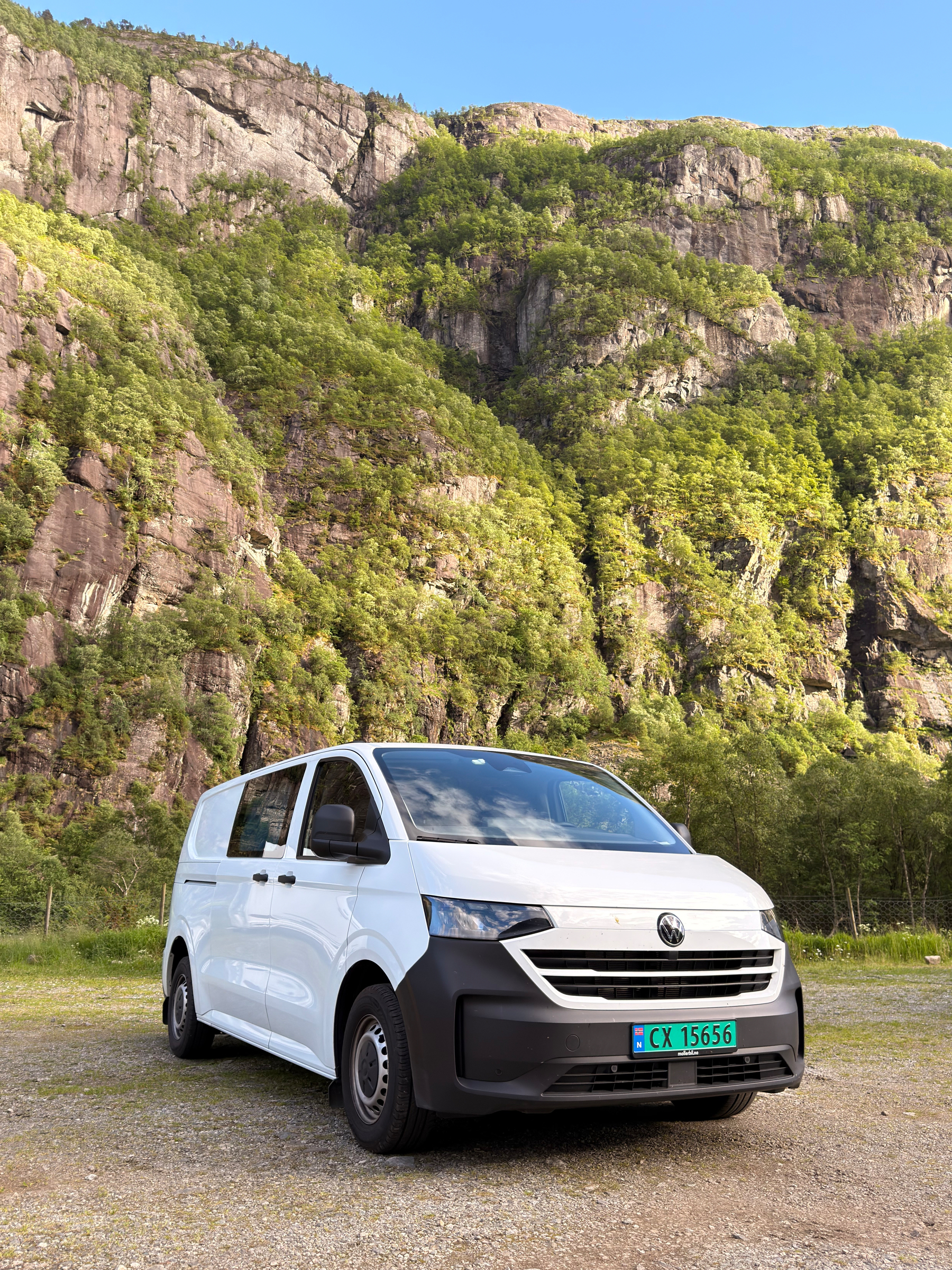 A white Volkswagen van is parked on a gravel area in front of a steep, green-covered rocky hillside under a clear blue sky.