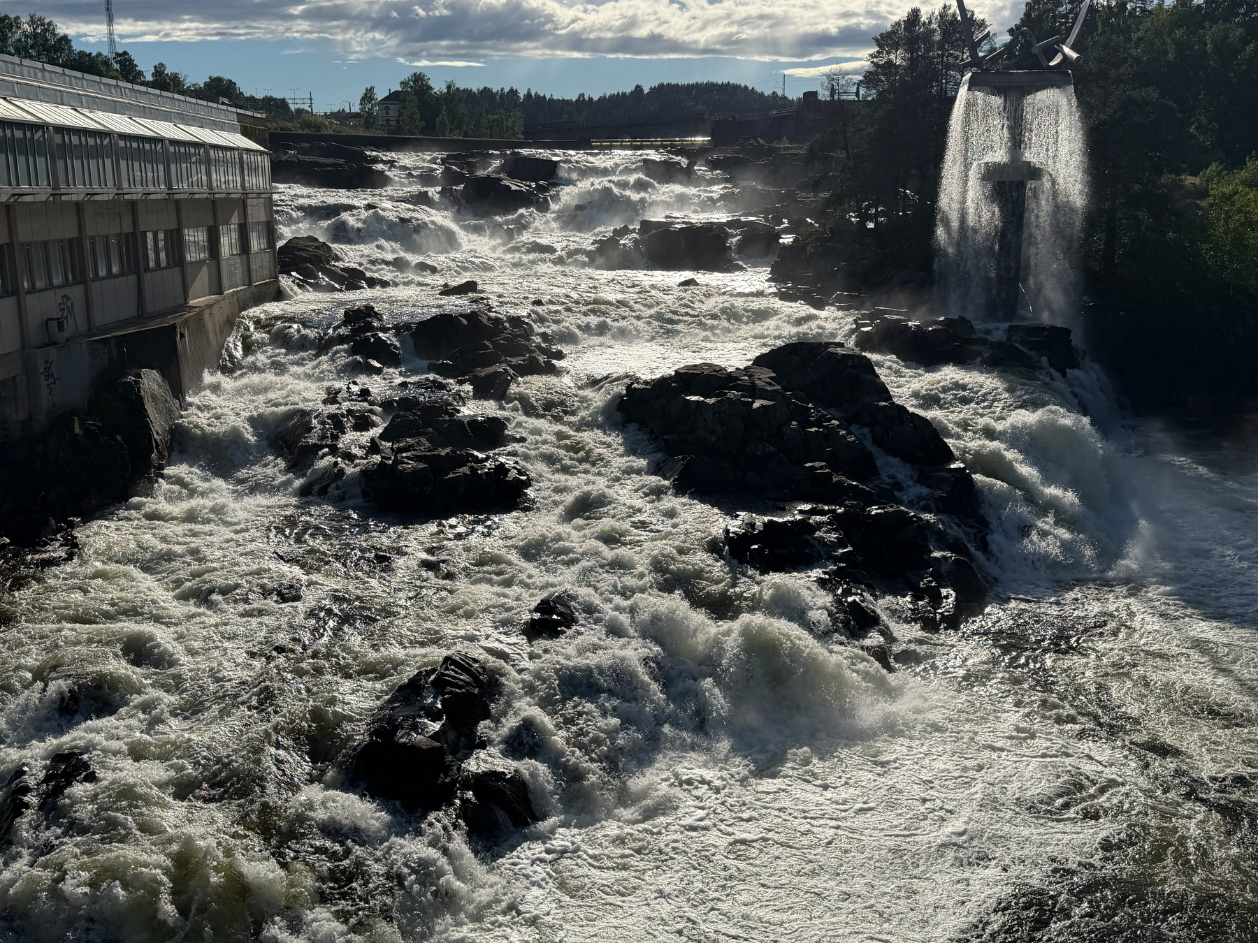 A powerful waterfall flows through rocky terrain beside a building and an elevated water channel, creating a dynamic and turbulent scene.