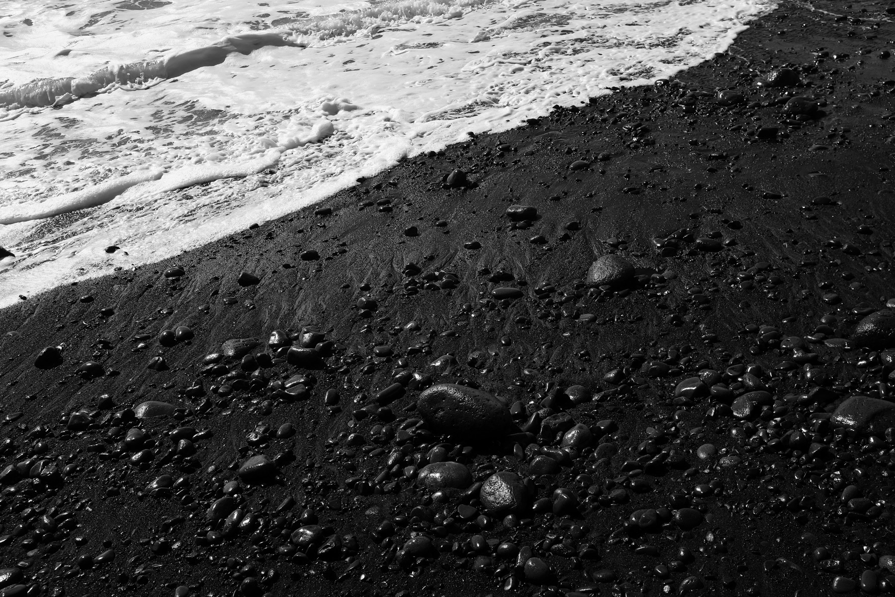 Black sand beach with scattered smooth stones, where waves gently wash ashore.