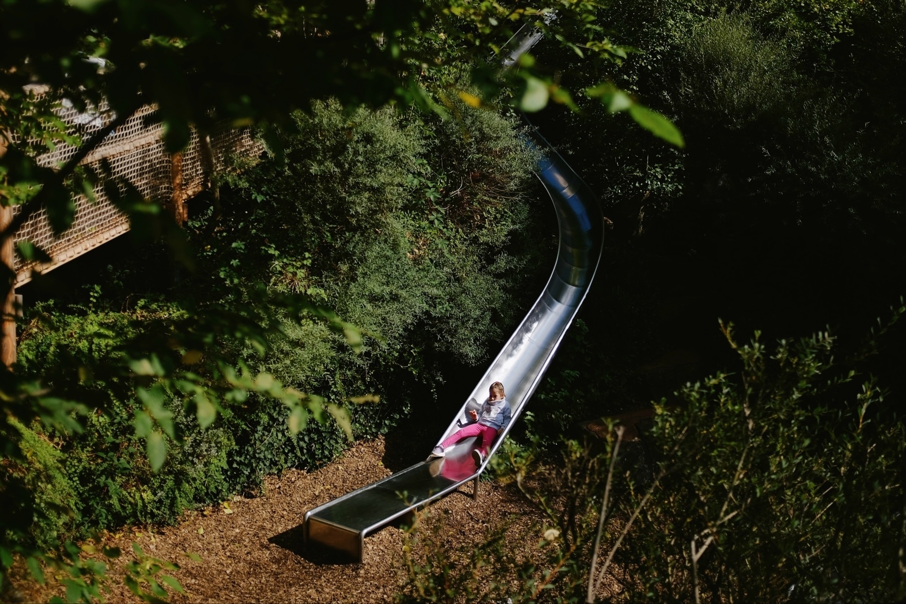 A child is sliding down a long, metal slide surrounded by lush green foliage and trees. The area is shaded, and there is a wooden walkway visible in the background. The child appears to be enjoying the slide.