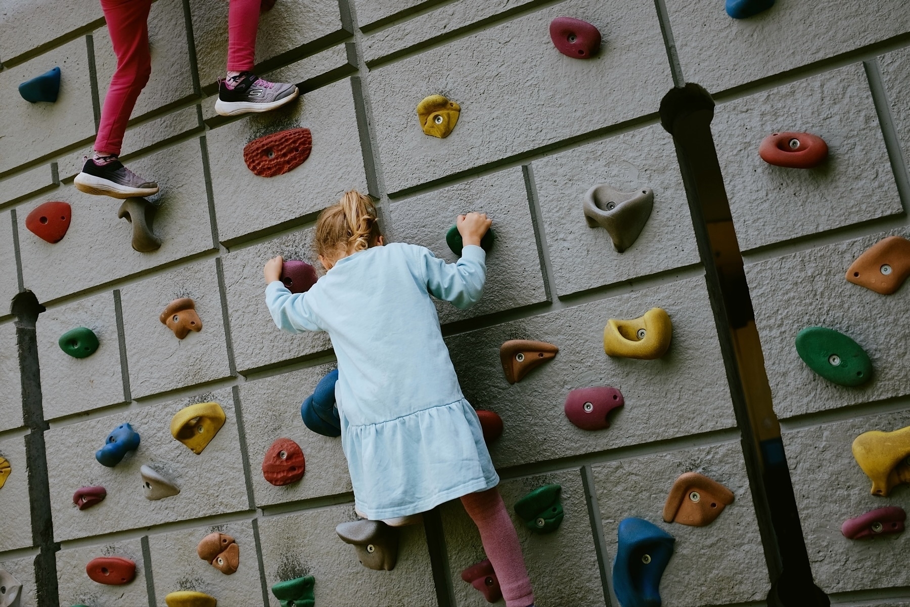 Two children are climbing an outdoor rock climbing wall with colorful holds. Only the legs of one child and the back of the other child are visible. The children are wearing casual clothing, including a light blue dress and pink leggings.