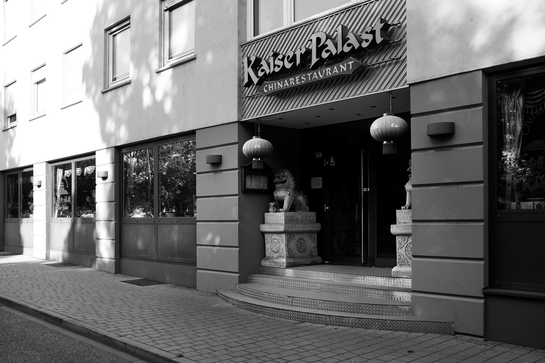 The image shows the exterior of “Kaiser Palast China Restaurant.” The entrance has decorative elements including traditional Chinese stone lion statues and hanging red lanterns. The building is modern with large windows.