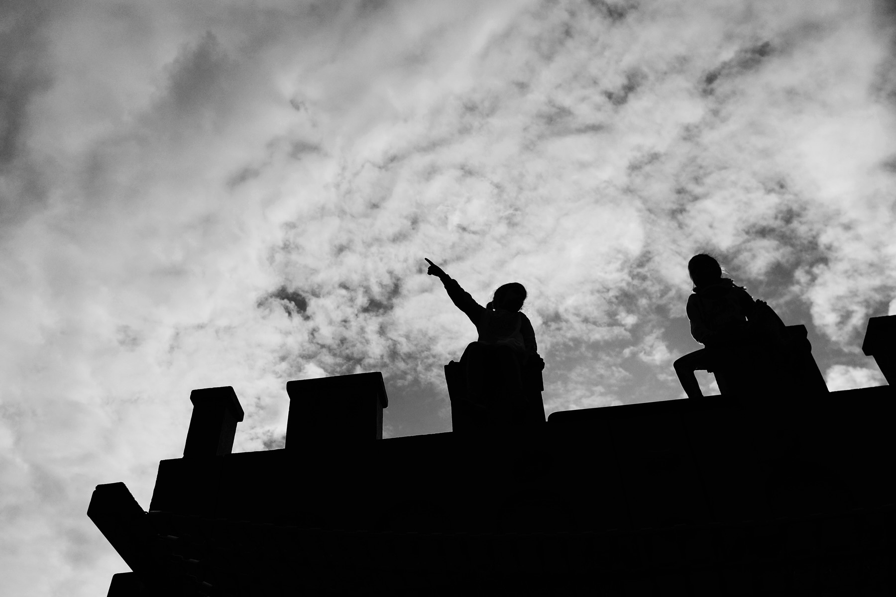 Silhouettes of two children sitting atop a fortress against a partly cloudy sky, with one of them pointing upwards.