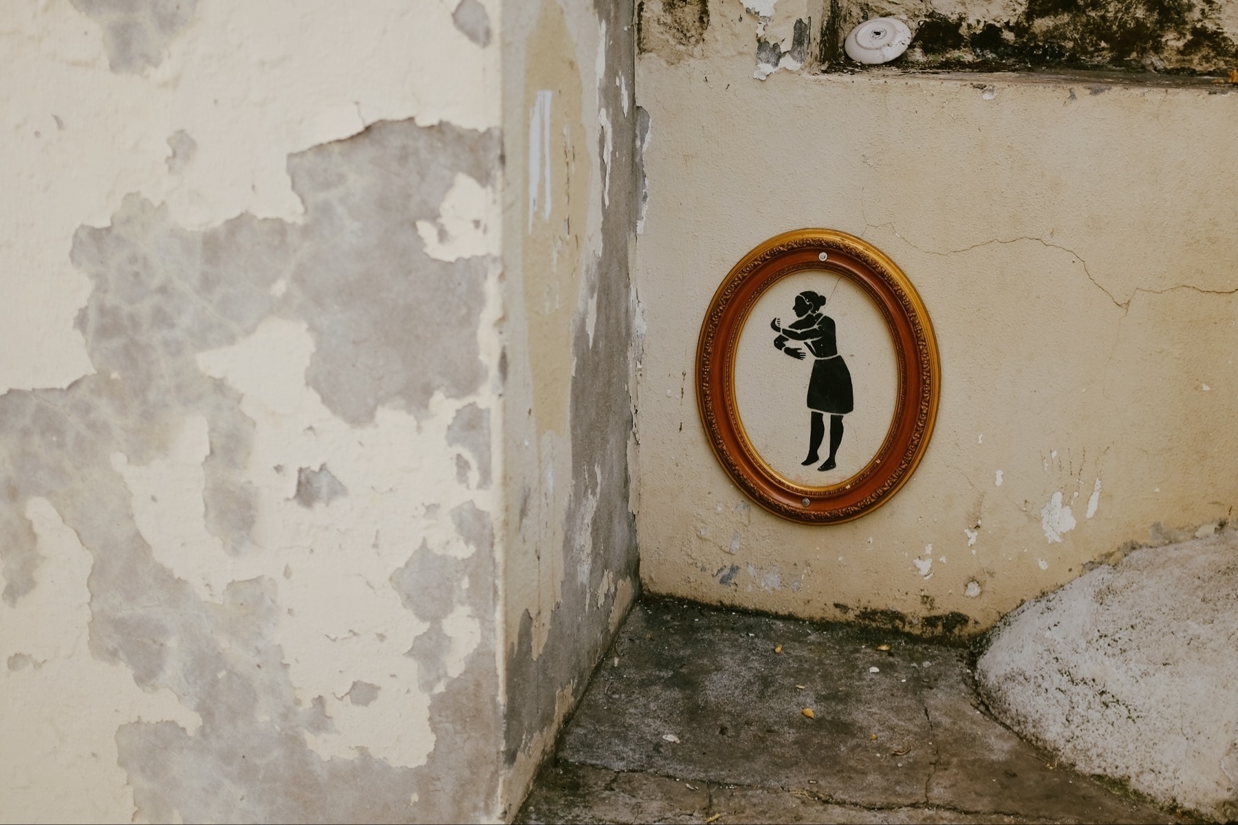 A weathered wall with peeling paint features a framed silhouette artwork of a person giving a hug. The frame is ornate and the setting has a rustic, aged look.