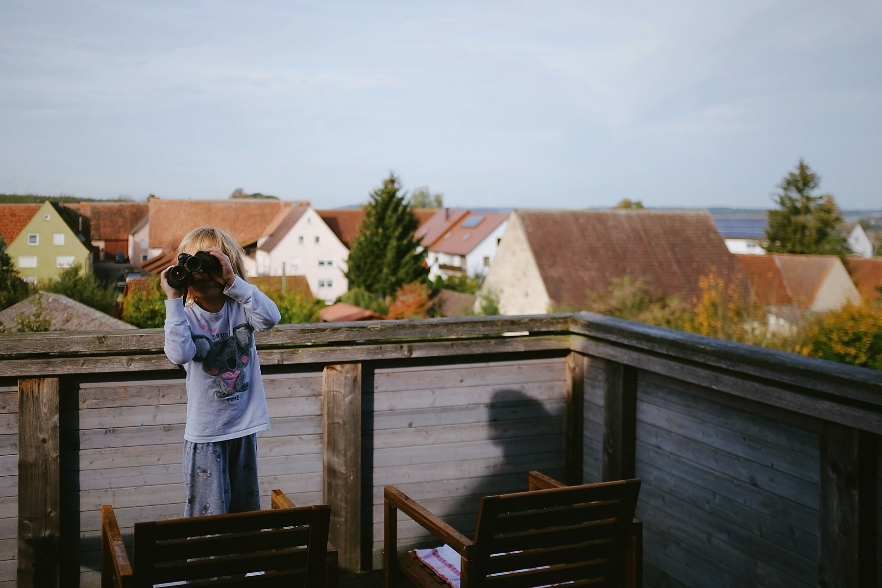 Child standing on a wooden chair on a balcony, looking through binoculars. Background features a village with houses and trees.