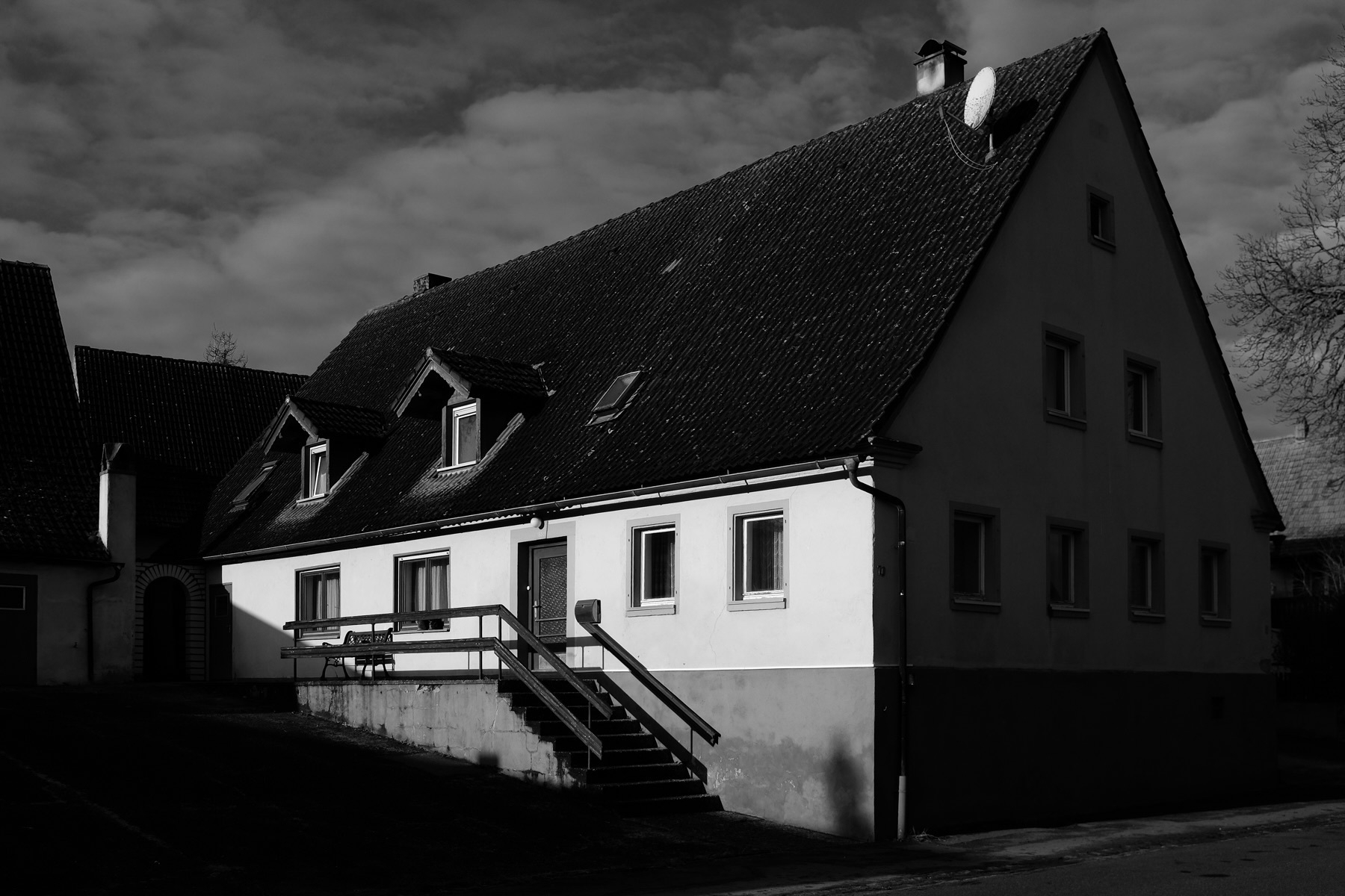 A traditional gabled house with small windows and exterior steps is captured in a dramatic black and white setting under a cloudy sky.