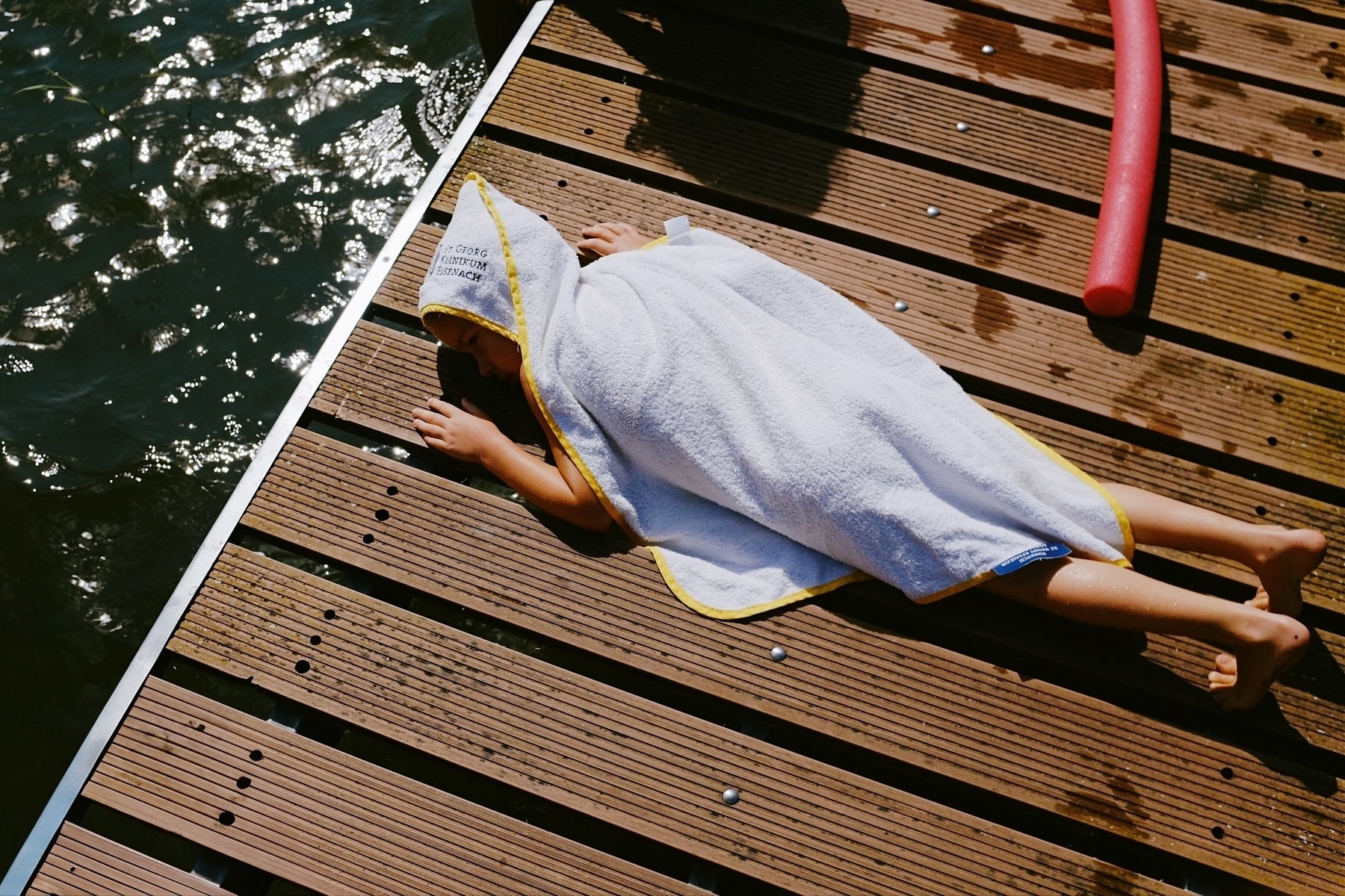 Child in a towel lying on a wooden dock beside water, post-swim relaxation.