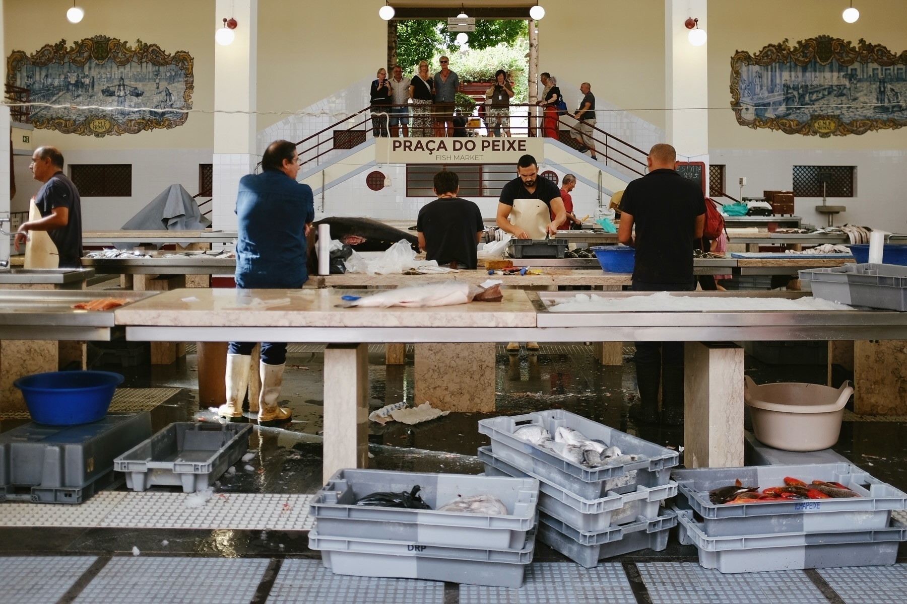 A fish market with several people working at large tables, preparing and displaying fish. There are stacks of gray plastic crates filled with fish. The market has a tiled floor and decorative murals on the walls. A sign reads “PRAÇA DO PEIXE”.