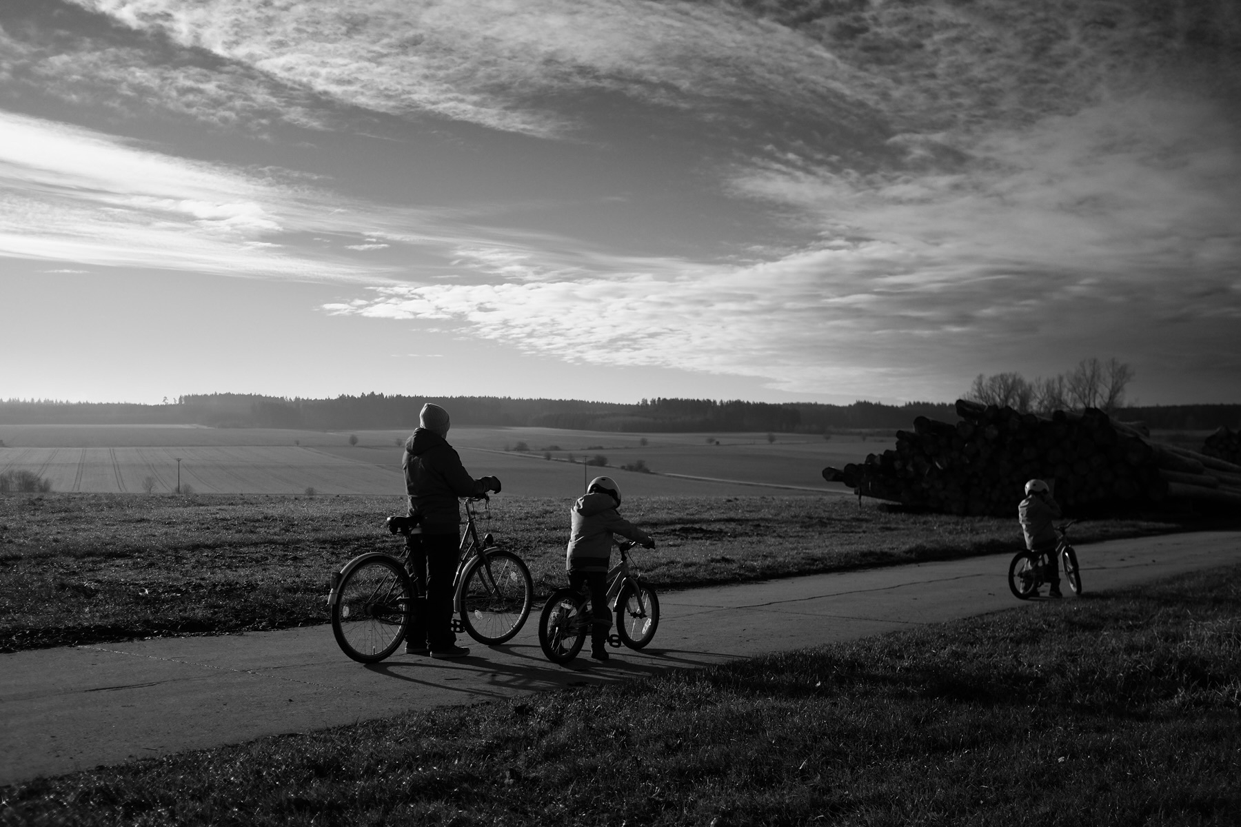 A group of three people, one adult and two children, are riding bicycles along a countryside path under a cloudy sky.