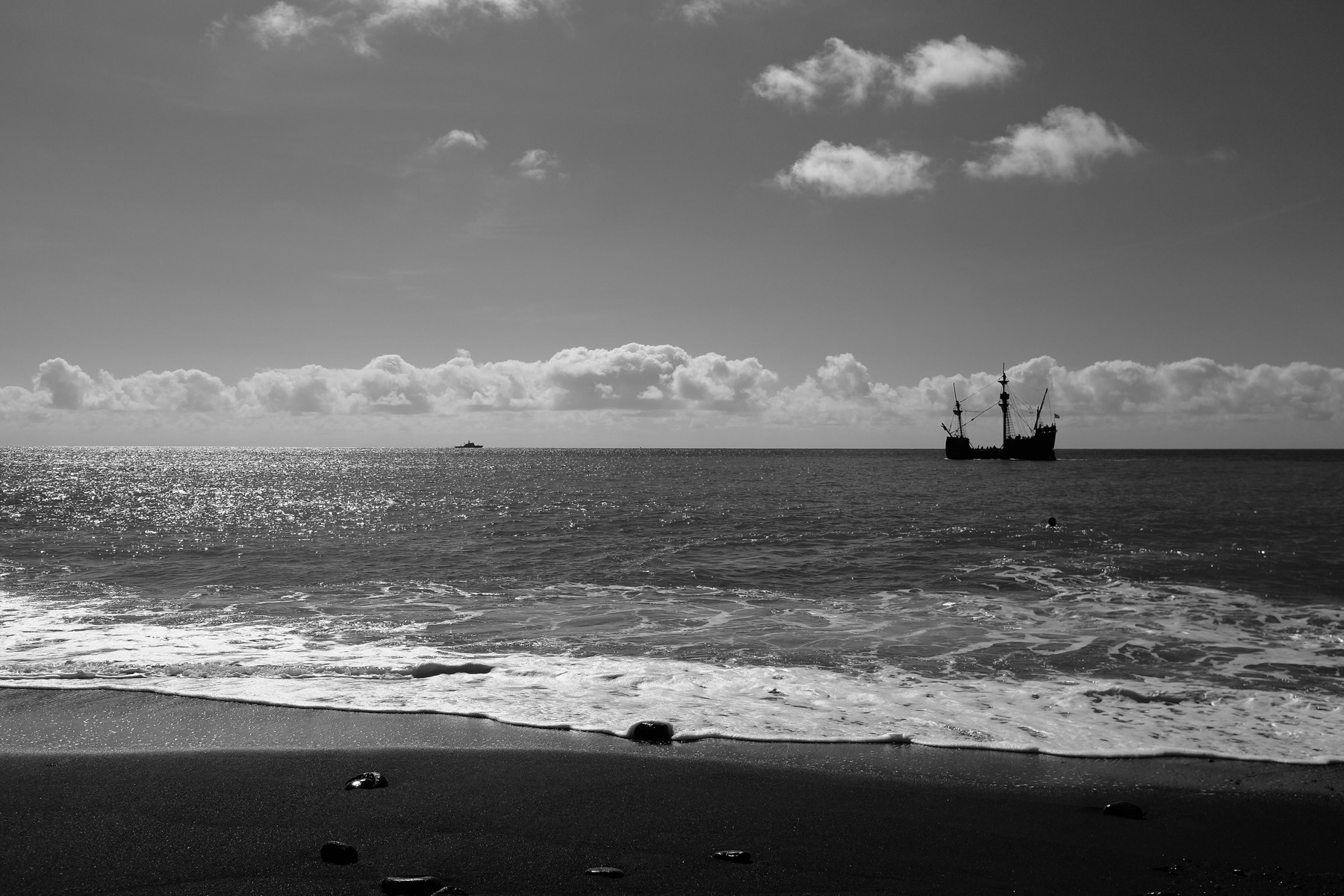 A black and white photo of a beach with gentle waves lapping the shore. In the distance, there’s a tall ship with multiple masts on the ocean, under a sky with scattered clouds. The sunlight shimmers on the water’s surface.