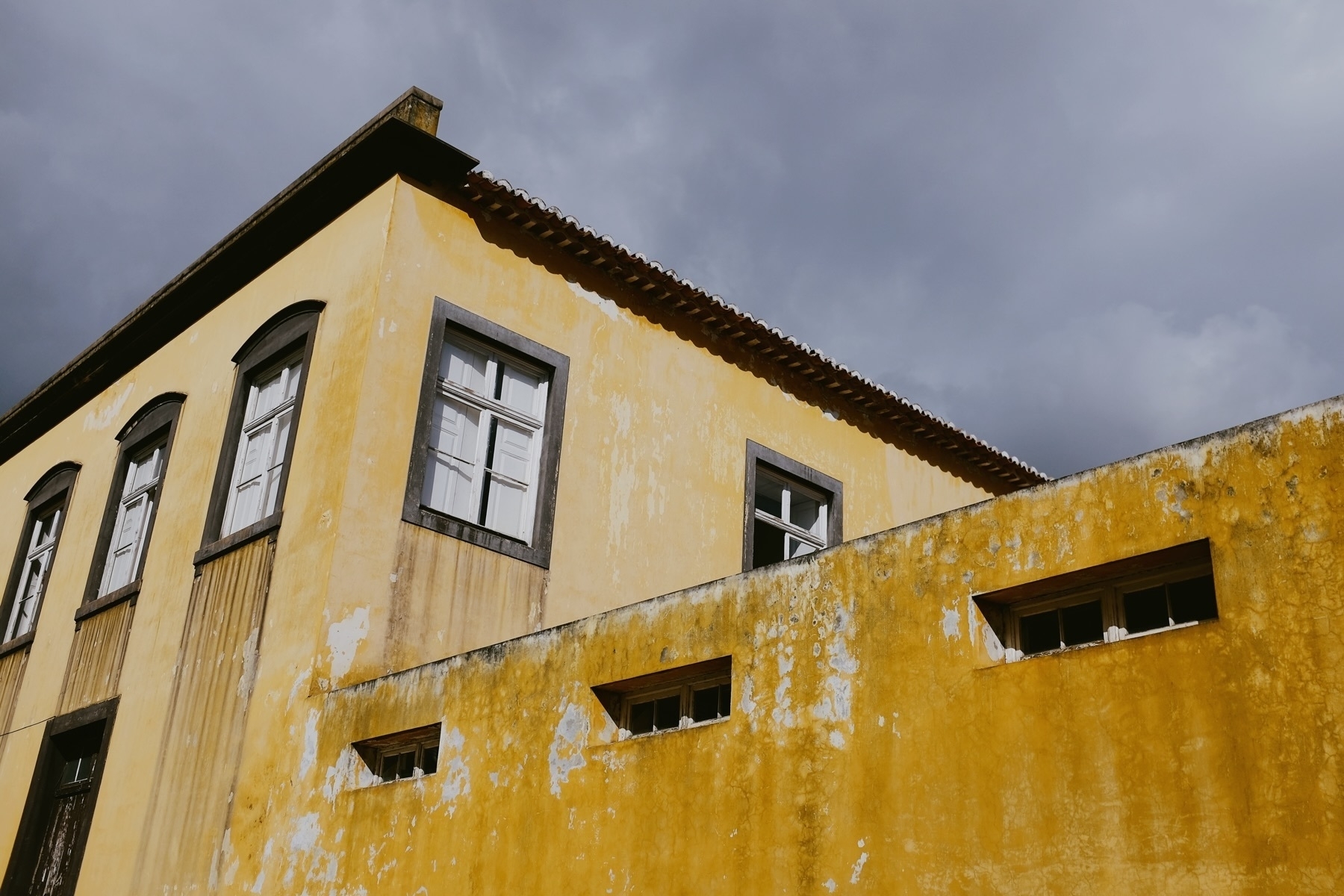 A yellow, weathered building with white-framed windows and a tiled roof against a cloudy sky.