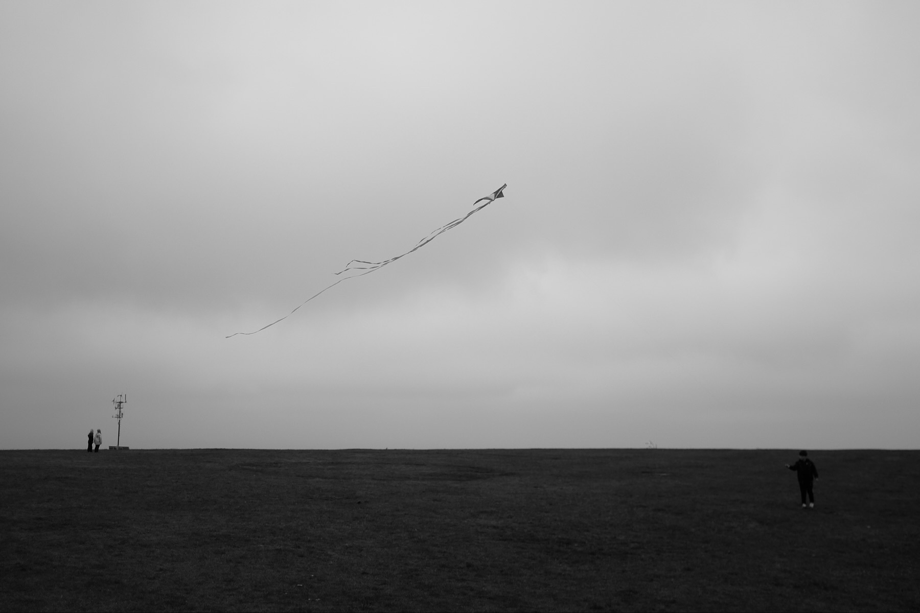 A black and white photo of a person flying a kite in an open field under a cloudy sky. In the background, there are two people standing near a weather station. The scene conveys a sense of solitude and open space.