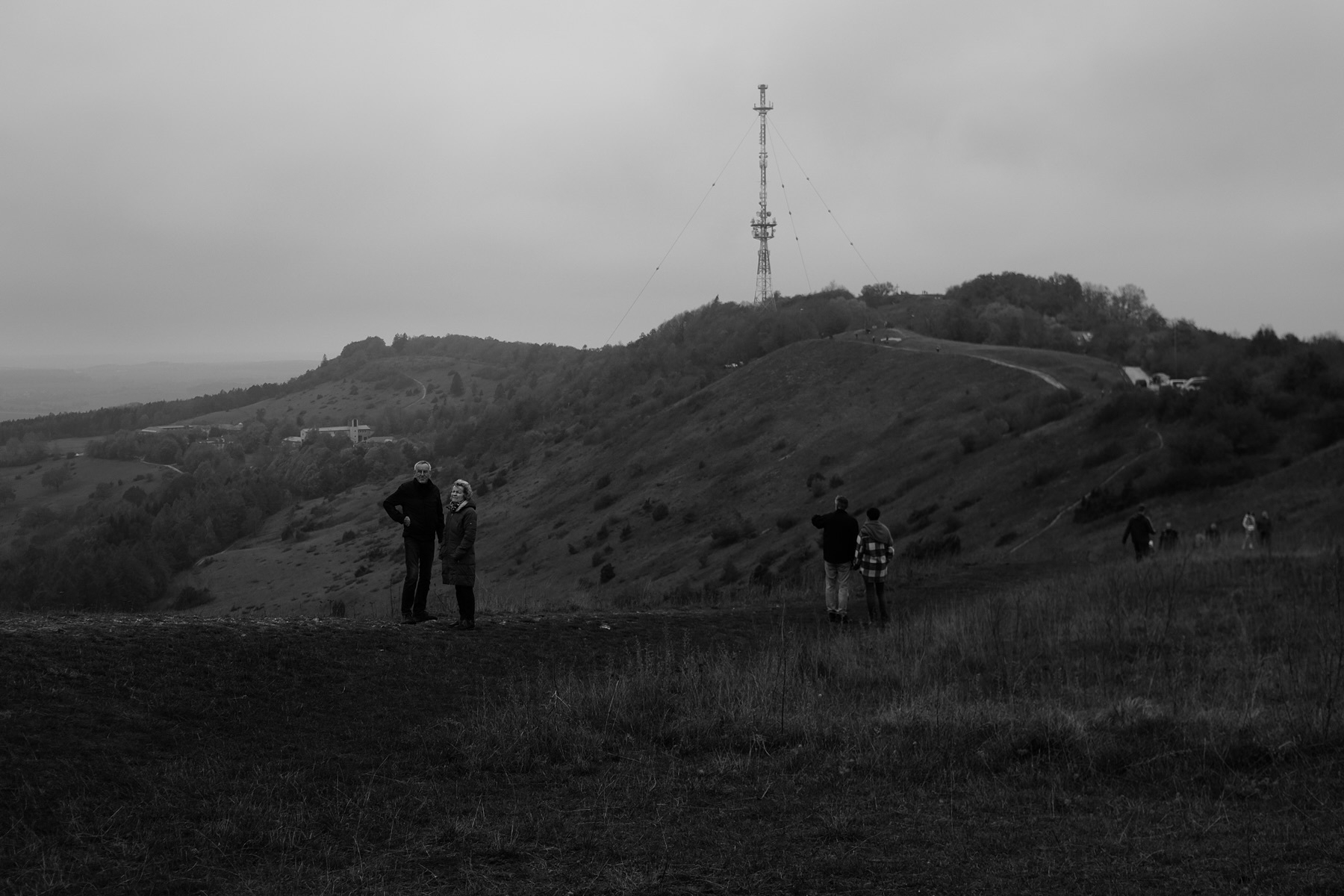 A black and white photo of a hilly landscape with a tall radio tower on a hilltop. People are scattered across the scene, some standing and others walking. The sky is overcast, creating a moody atmosphere.