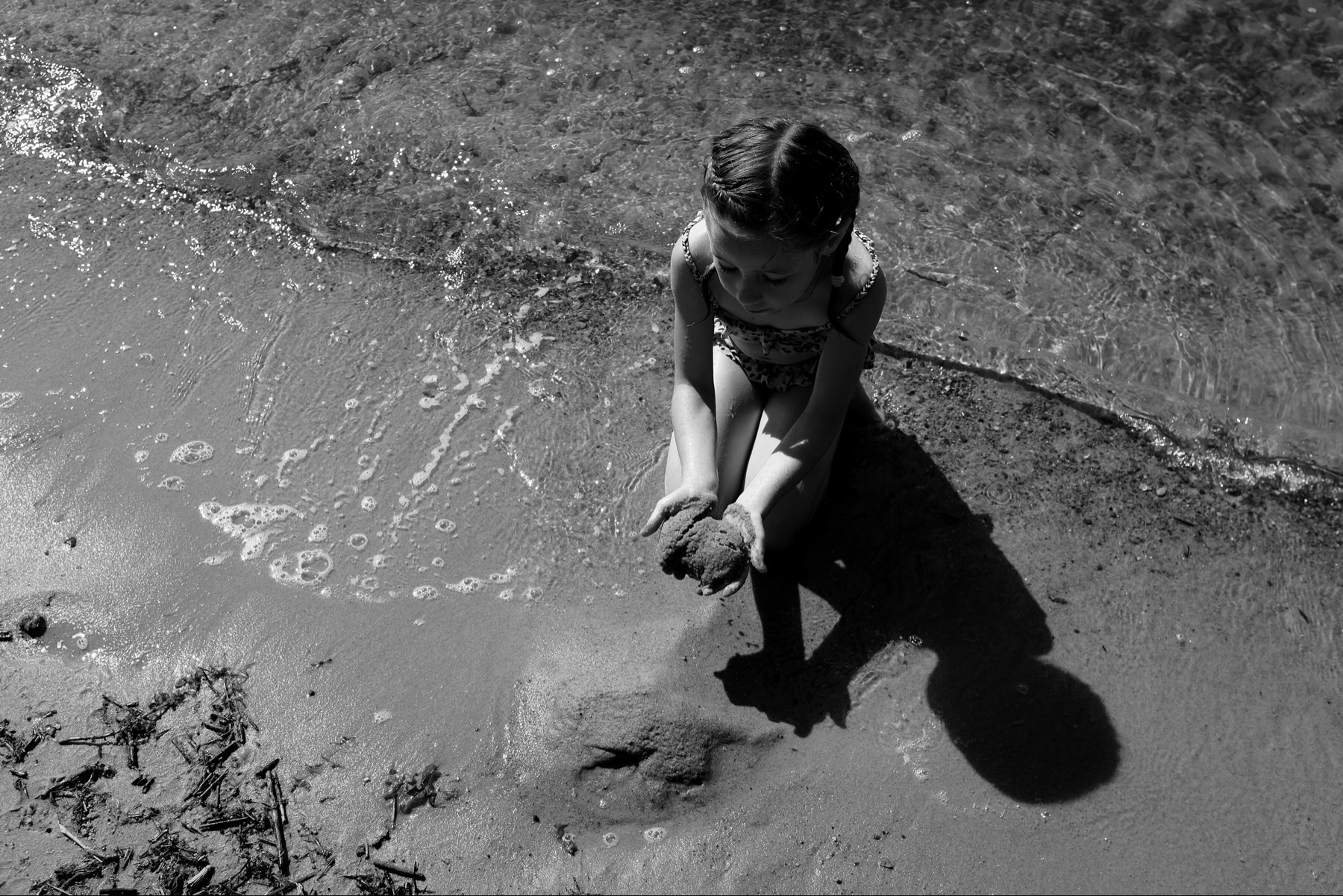 Child playing with sand at water’s edge, casting a shadow, in black and white.