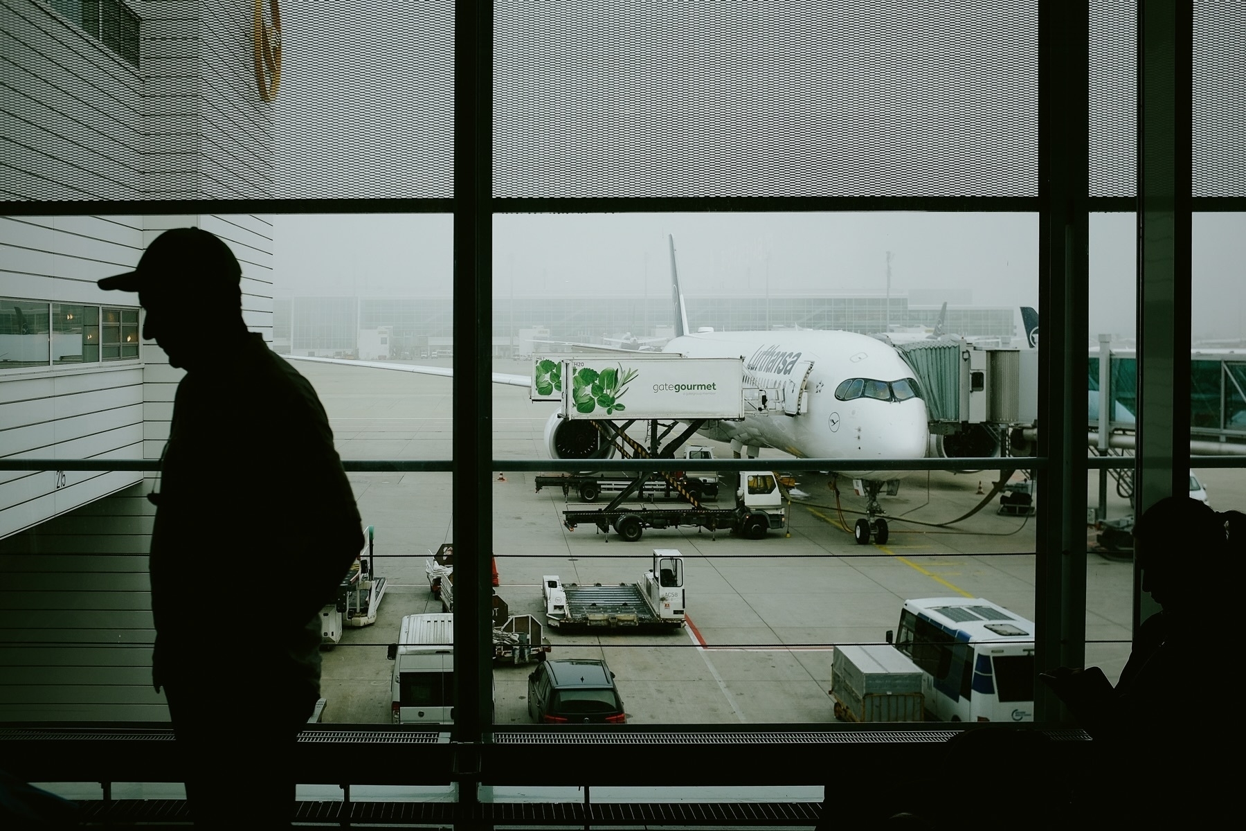 Silhouette of people inside an airport terminal, with a Lufthansa airplane visible on the tarmac outside. Ground vehicles and catering trucks are servicing the aircraft.