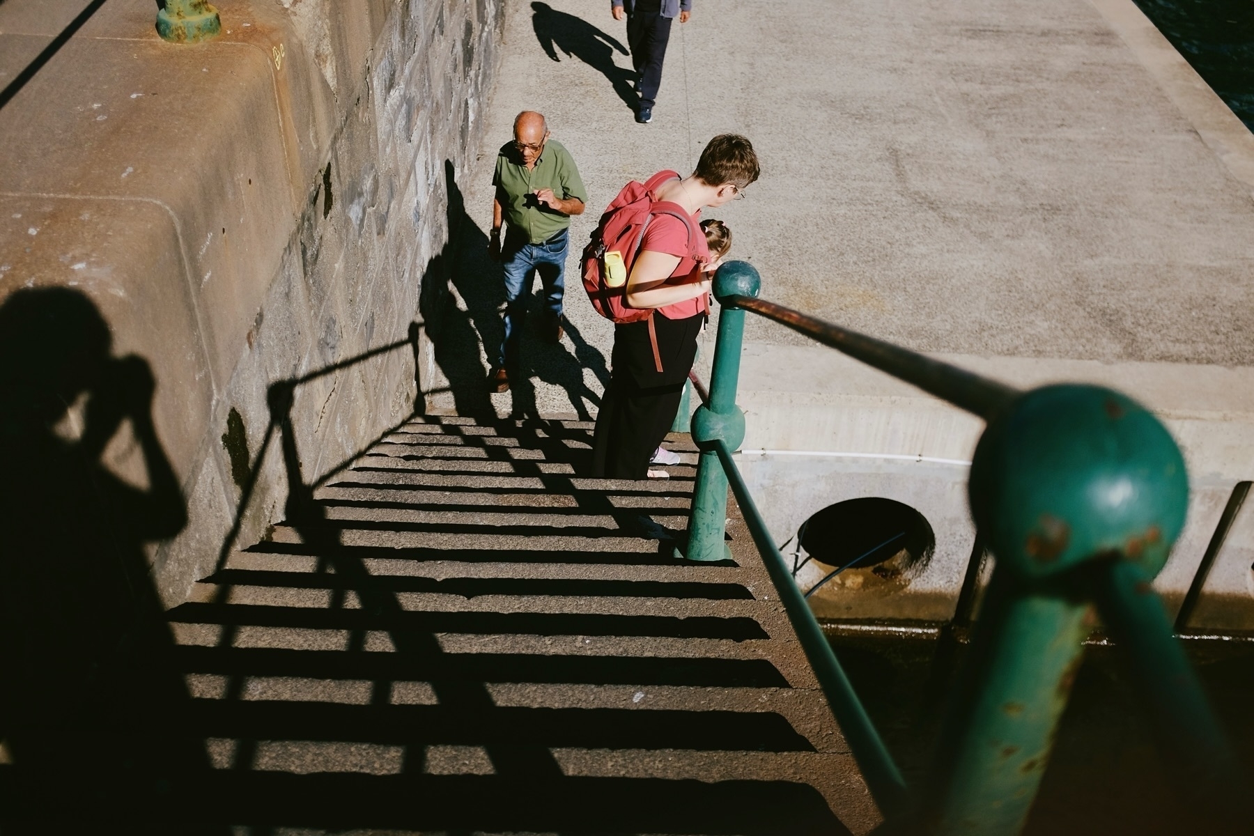 People descending a sunlit staircase near a stone wall. A person with a pink backpack is followed by another person, while shadows are cast on the steps. A green railing is in the foreground.