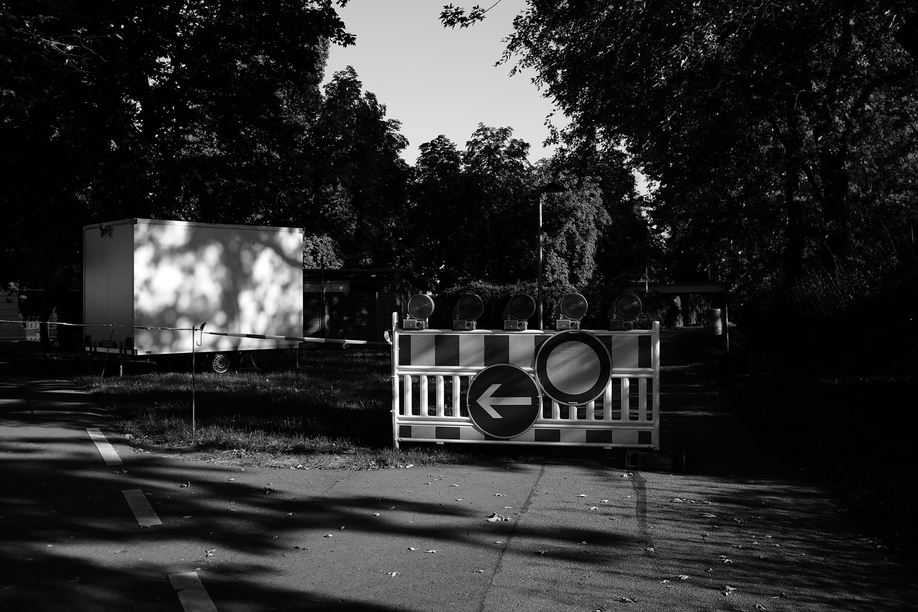 The image shows a temporary roadblock with a barrier and a detour sign indicating a left turn, set up on a pathway in a park-like area. Behind the barrier, a white trailer and trees can be seen.